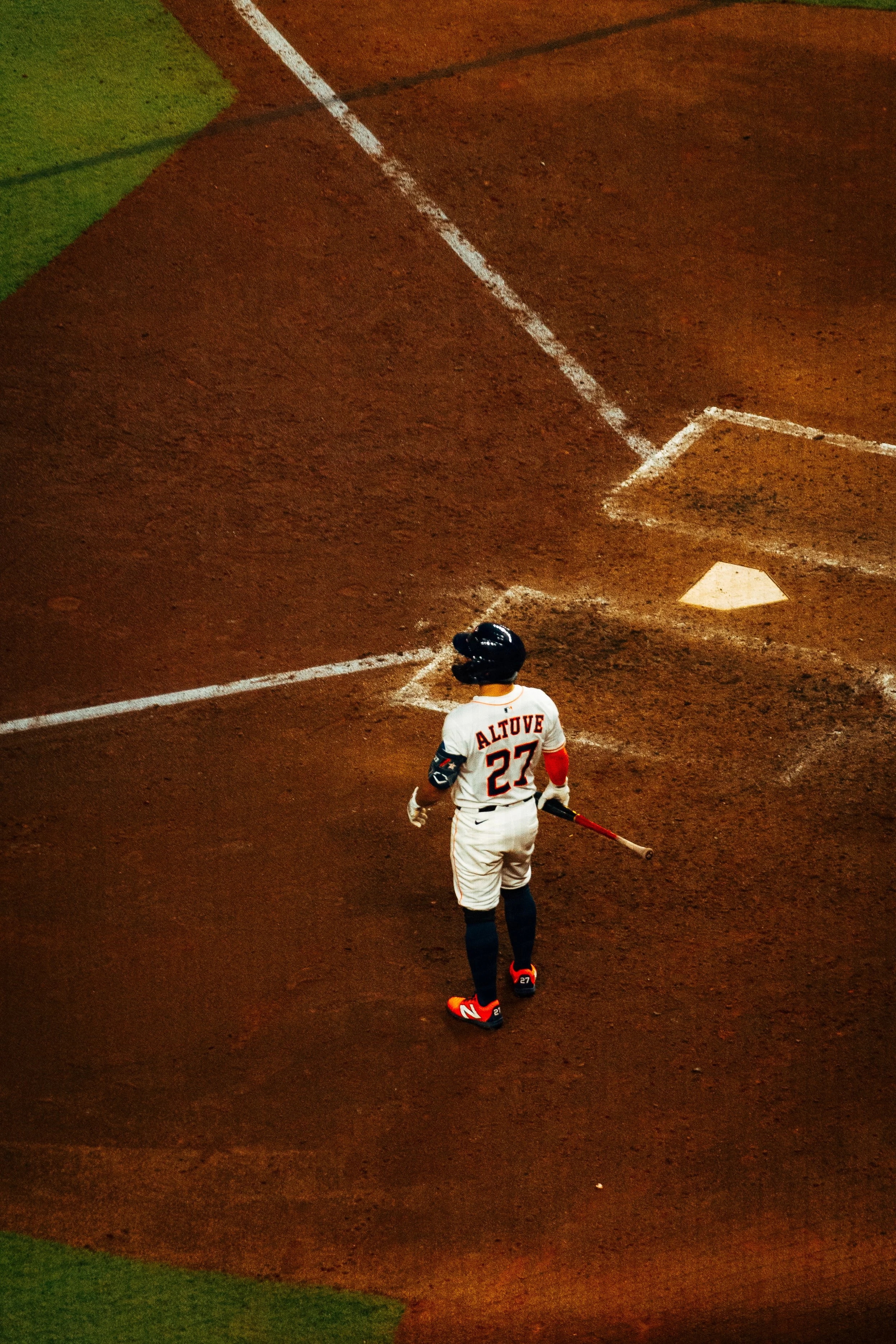 Baseball player with jersey number 27 walking on the field near home plate, holding a bat, with dirt and chalk lines visible.
