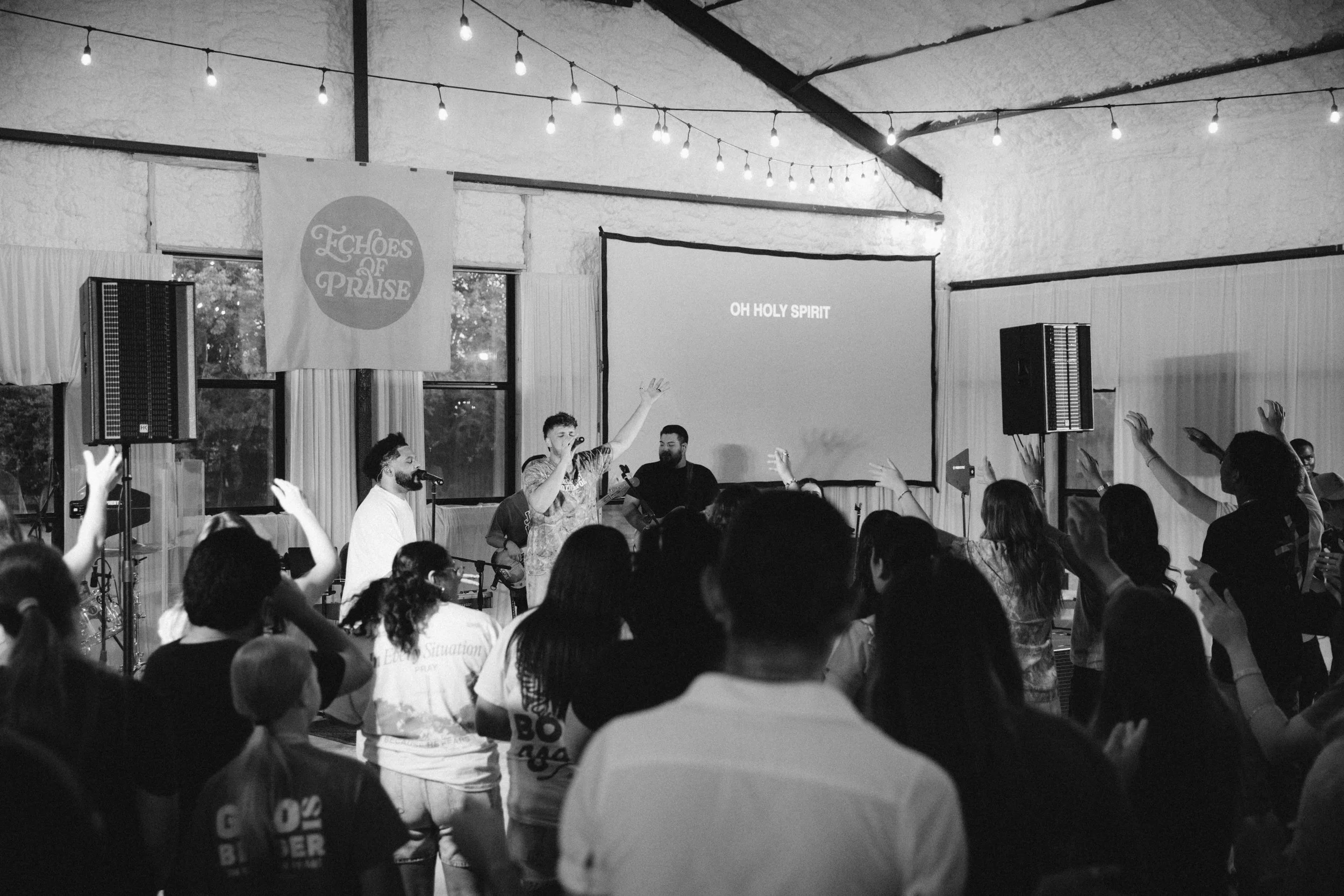 A black and white photo of a church gathering where people are standing, some with hands raised, during a worship session. A band and singers perform near a large screen that displays the words 'OH HOLY SPIRIT'. Lighting fixtures hang from the ceilin