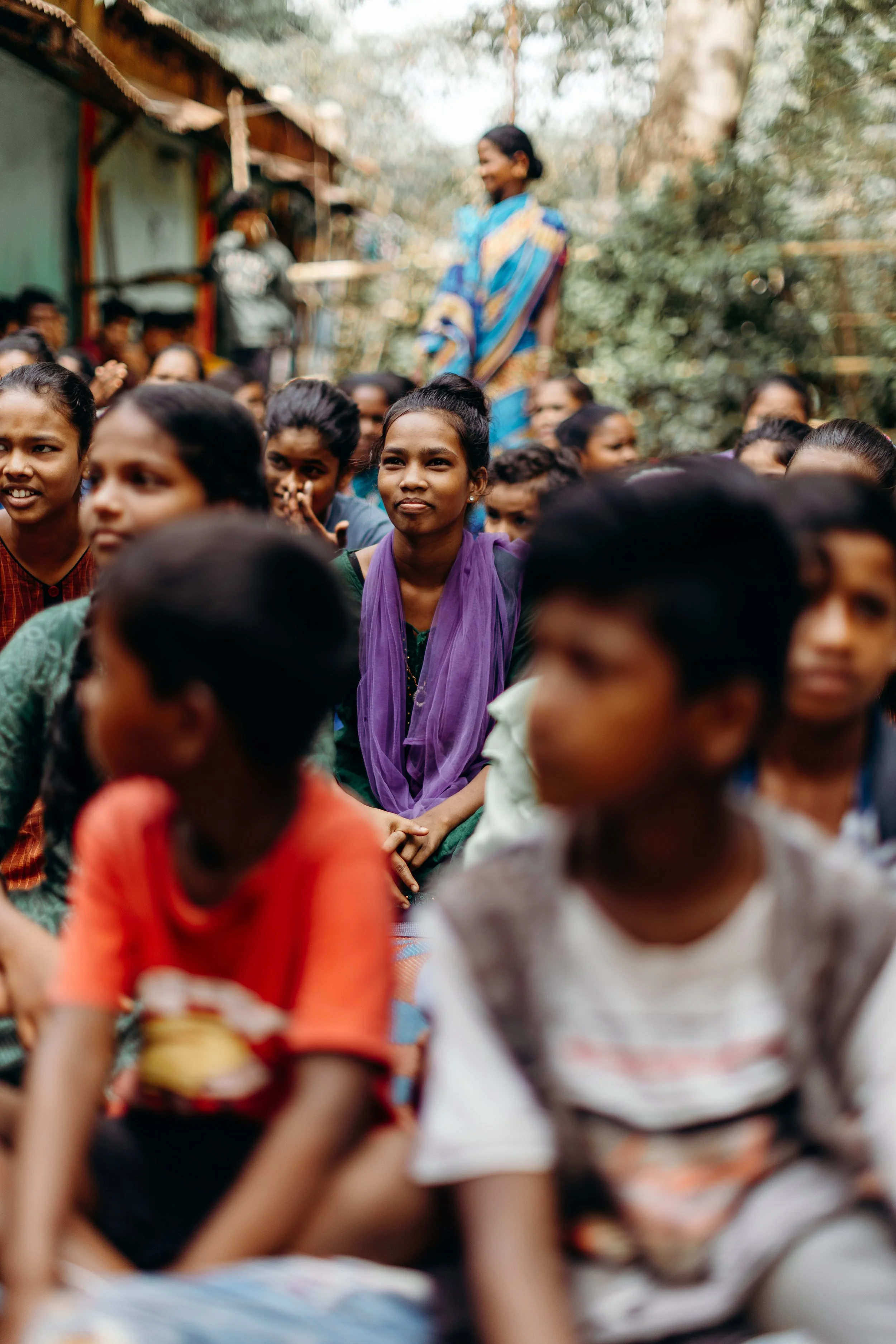 A group of children and a woman outdoors, some children sitting and some standing, with trees and a building in the background.