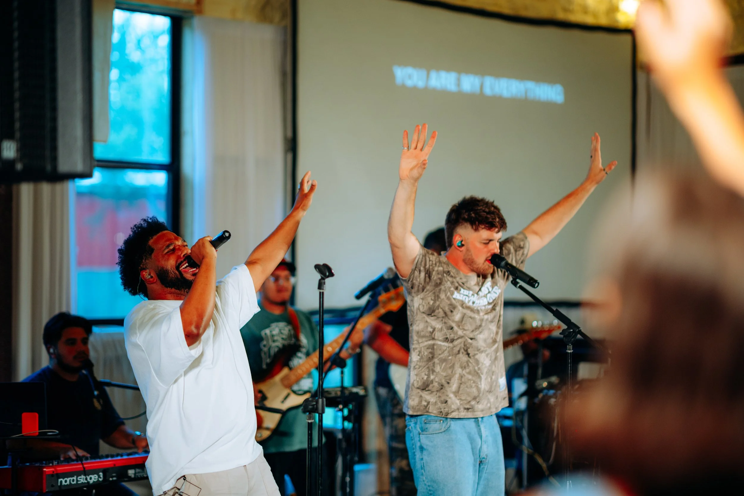 Two male singers with microphones perform with their hands raised, singing passionately, in front of a band at an indoor concert or worship event. A banner with the text 'YOU ARE MY EVERYTHING' is projected on a screen behind them.