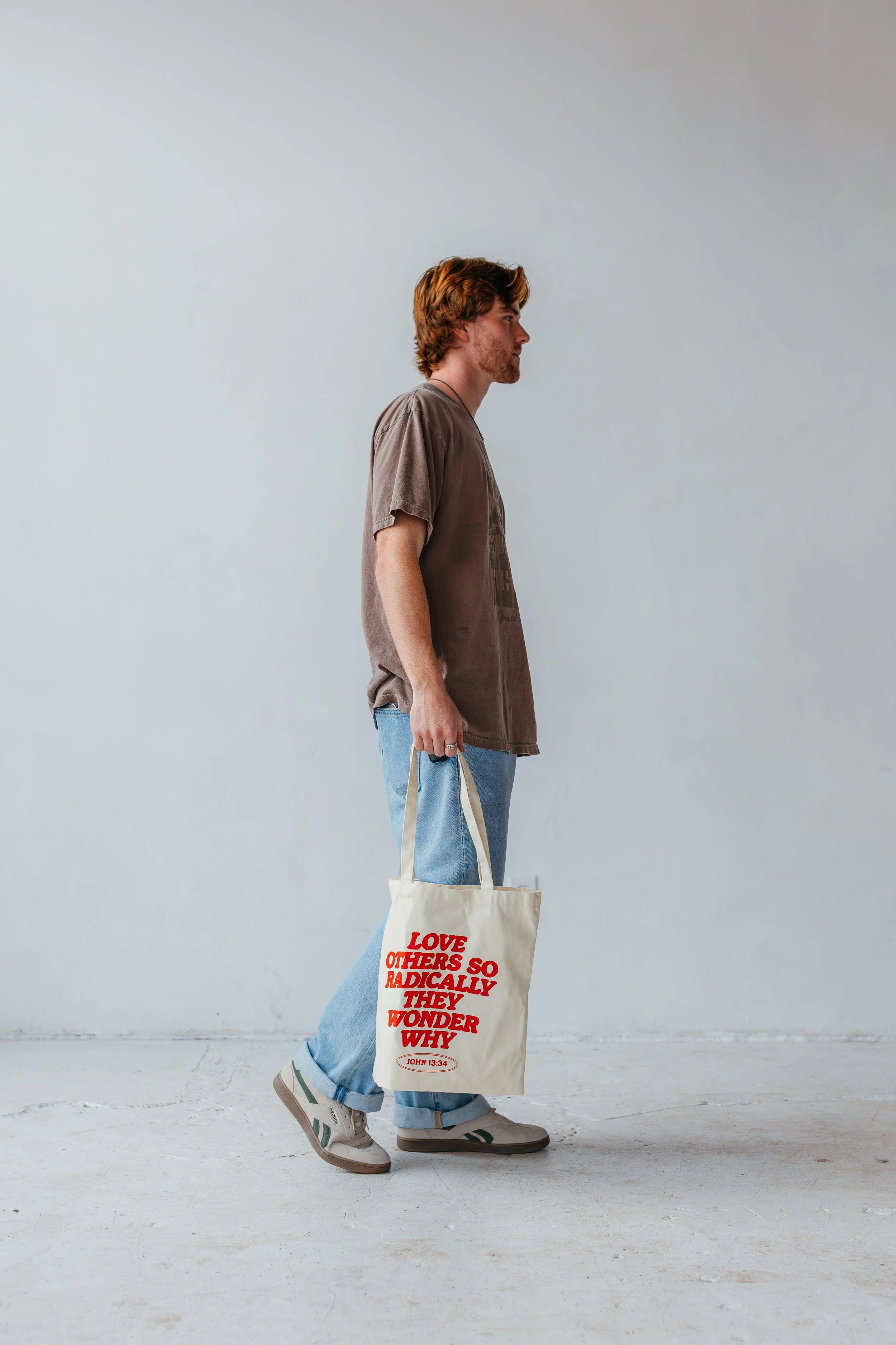 A young man with brown hair and a beard holding a beige tote bag with red text that reads "LOVE OTHERS SO RADIALLY THEY WONDER WHY," standing against a plain light gray wall. He is wearing a brown t-shirt, light blue jeans, and gray sneakers.