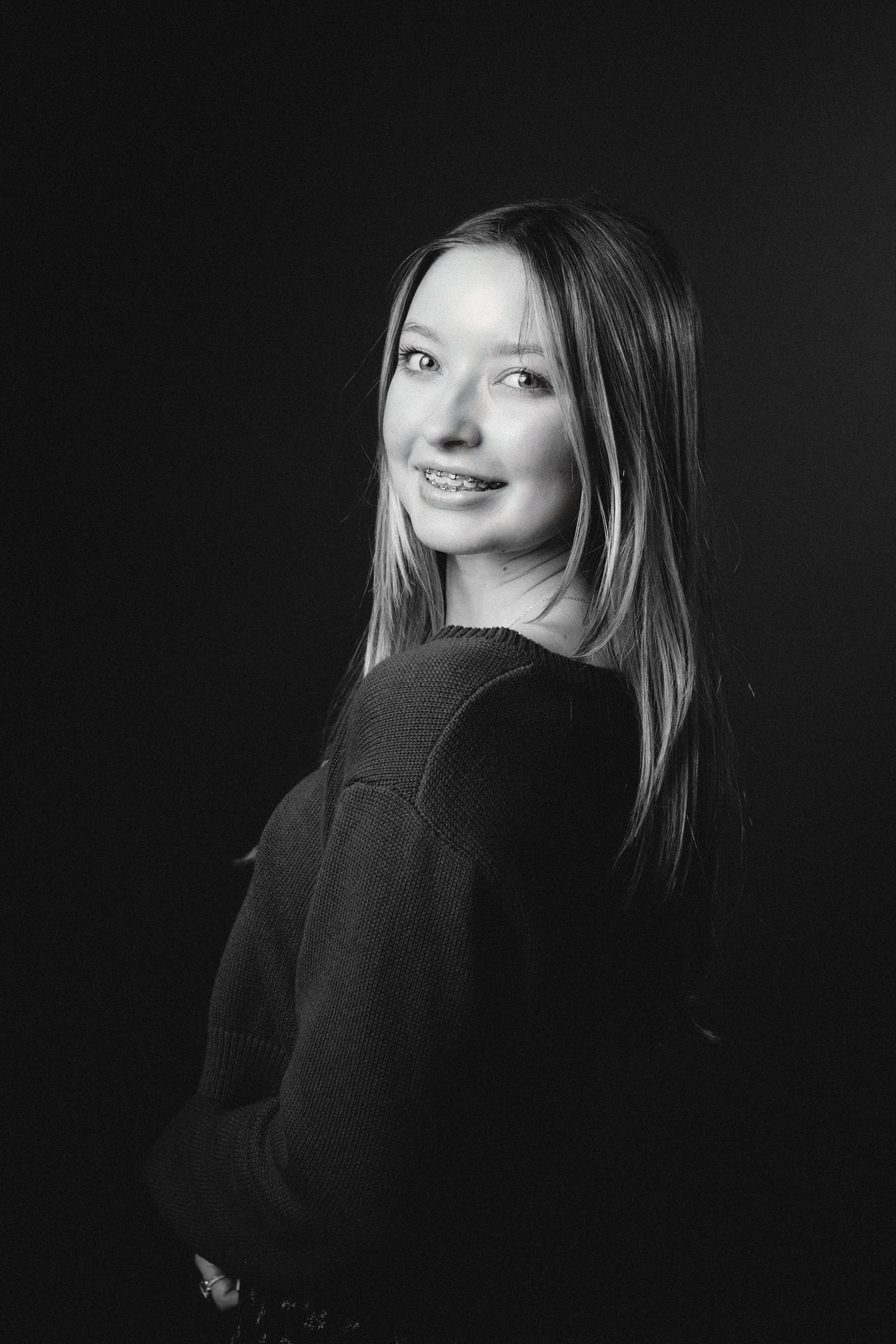 Black and white portrait of a young woman smiling and looking at the camera, wearing a dark sweater, with her hair down against a dark background.