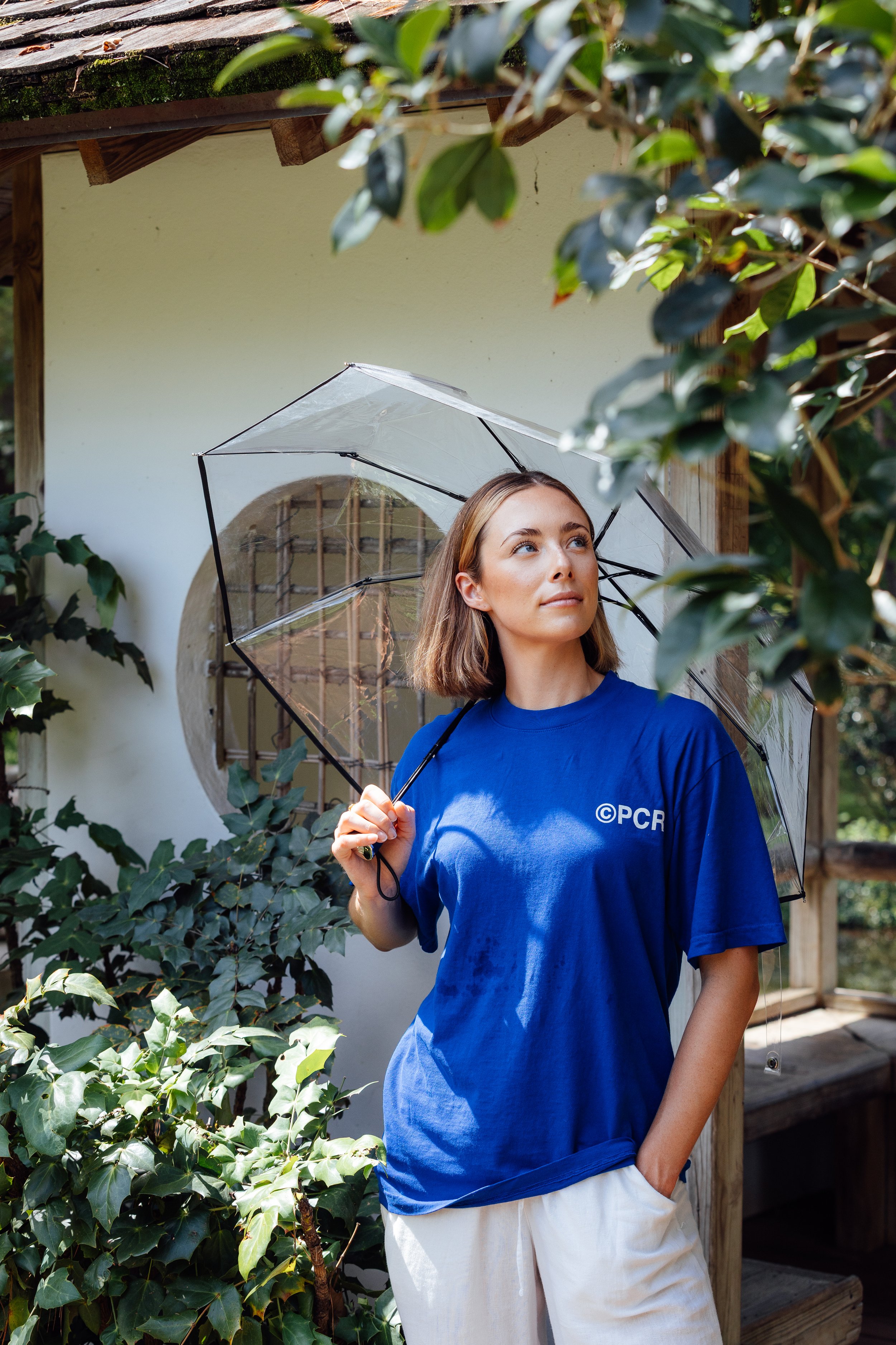 A woman in a blue T-shirt holding a clear umbrella, standing outdoors among green foliage near a white building, looking off into the distance.