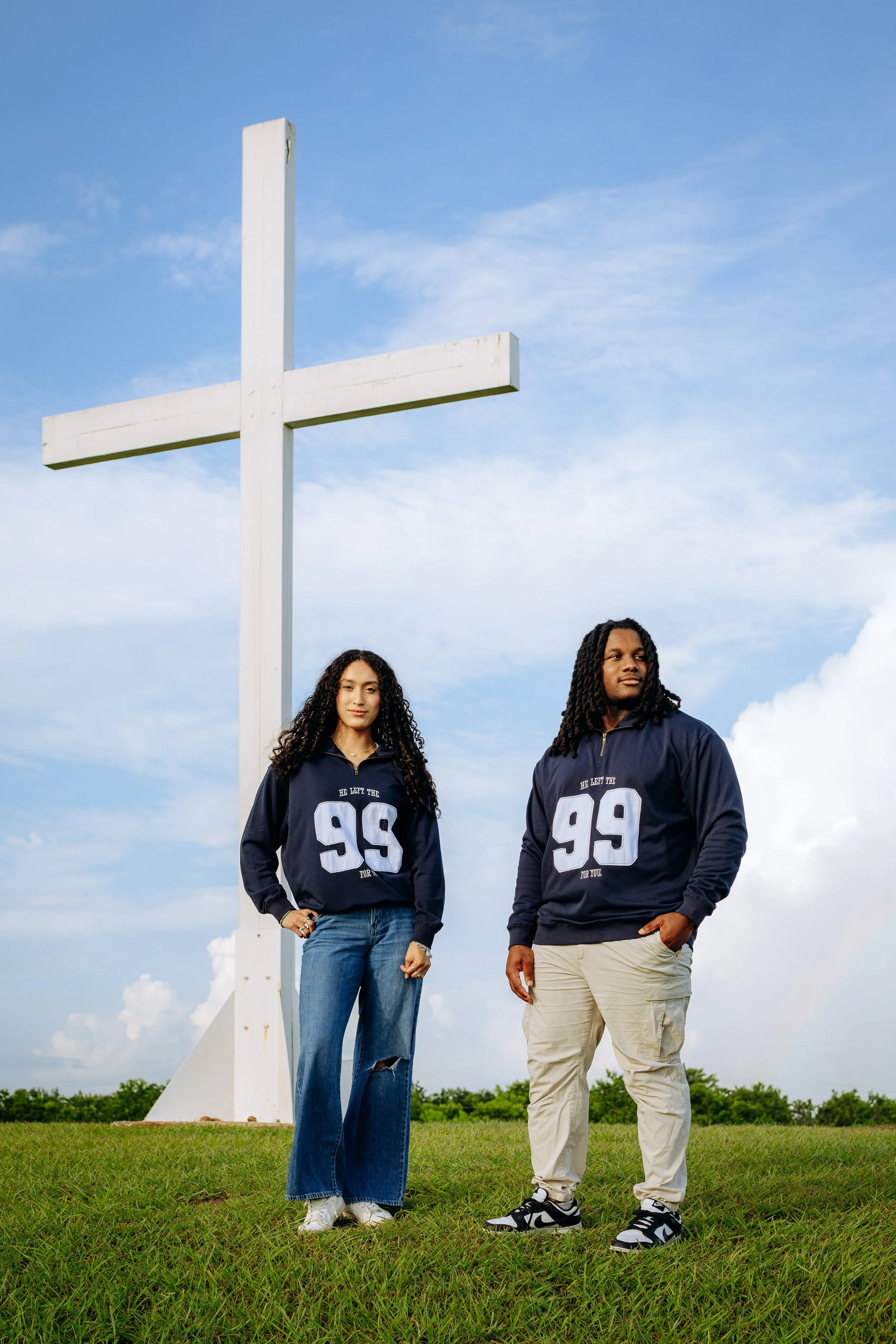 Two young adults standing on grass in front of a large white cross, both wearing navy sweatshirts with the number 99, under a bright blue sky with scattered clouds.