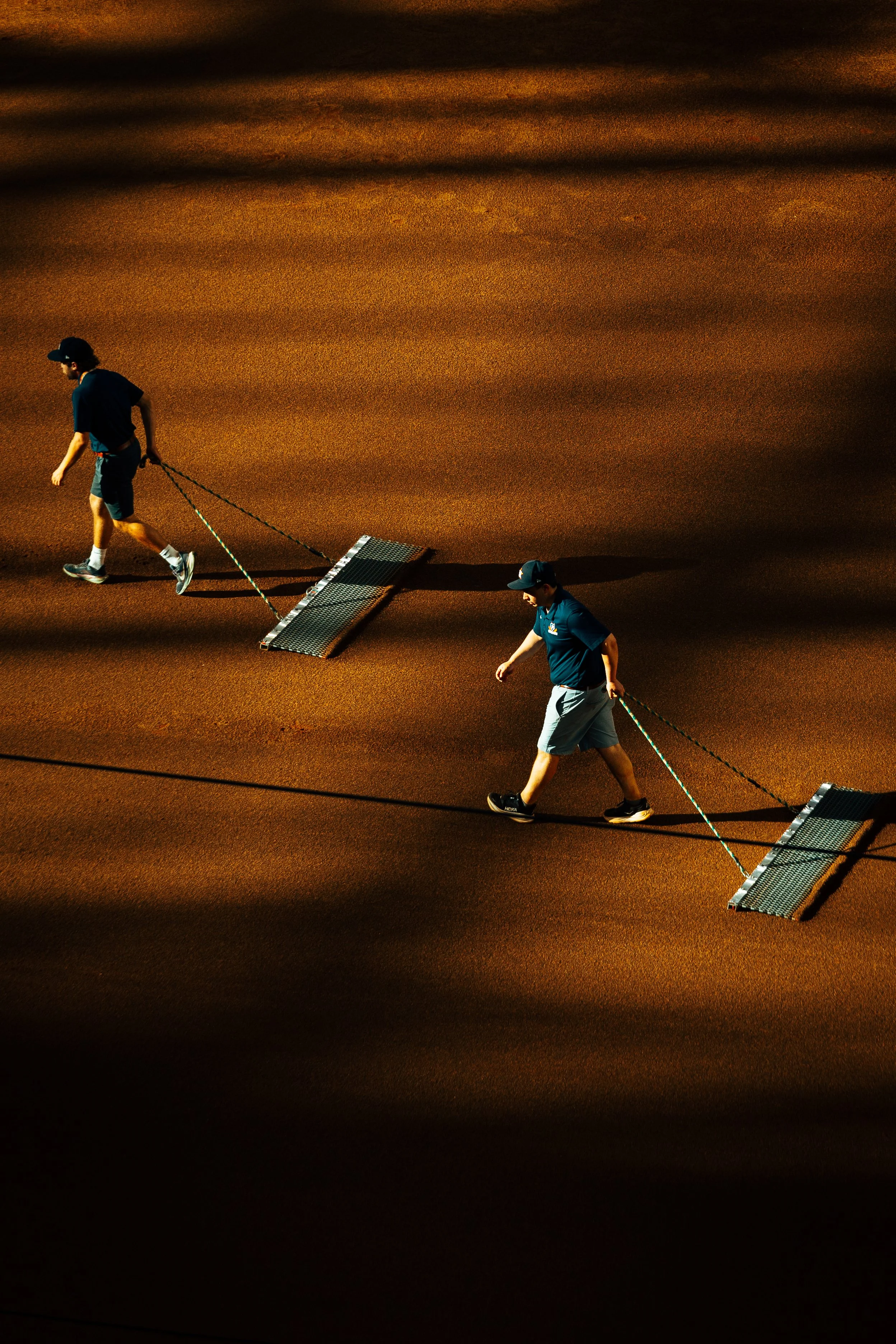 Two maintenance workers walking across a baseball field at night, pulling a roller behind them.