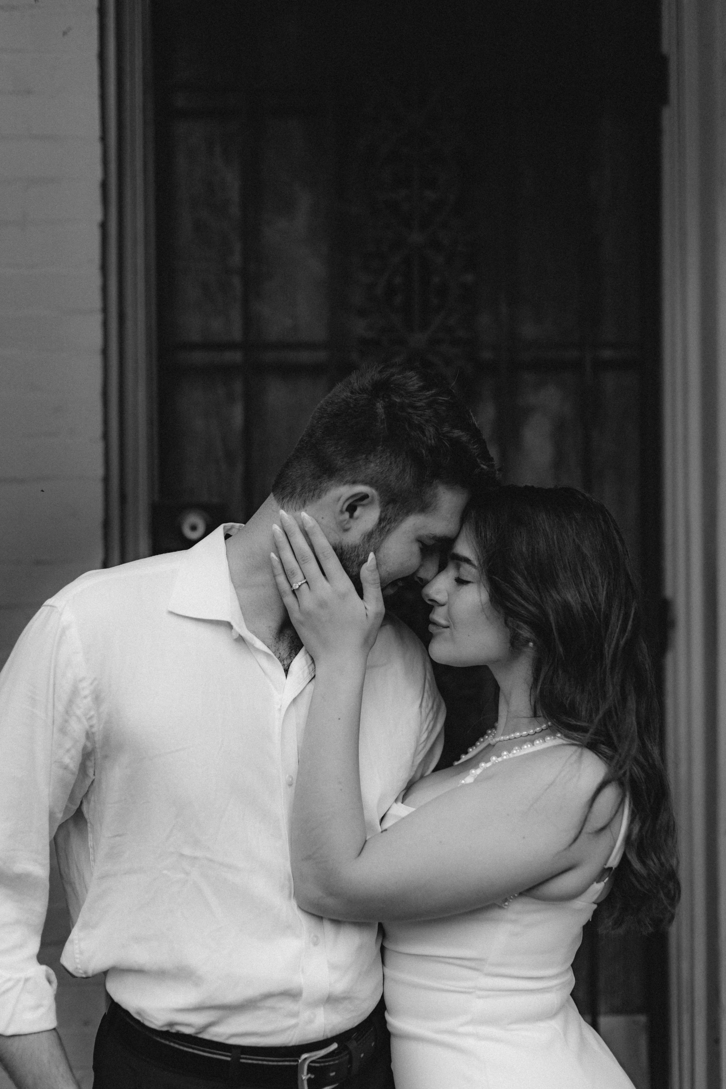 A black-and-white photo of a couple in an intimate moment, touching foreheads with eyes closed, standing close together indoors.