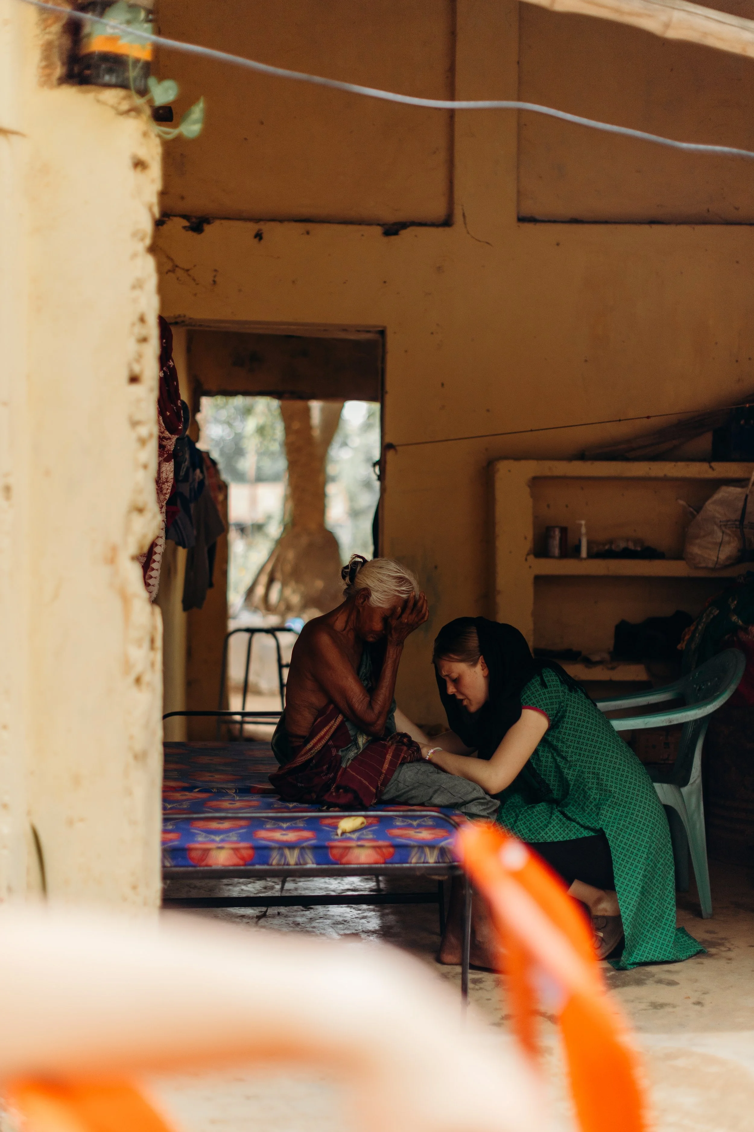 A younger woman is kneeling beside an older, shirtless man sitting on a bed, holding his hand and comforting him. The scene is inside a modest room with a simple bed and minimal furniture.