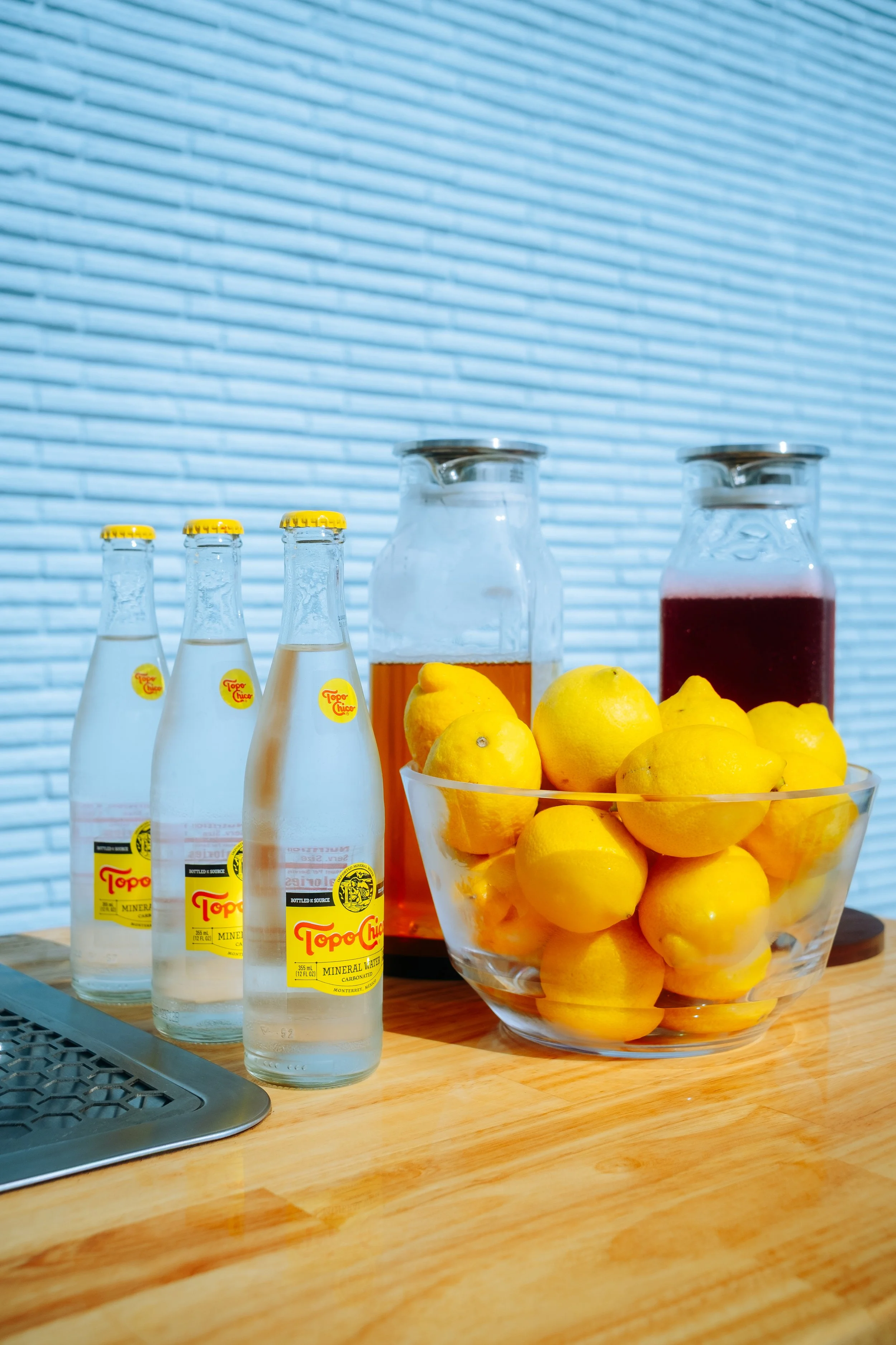 Three bottles of Topo Chico mineral water, a bowl of lemons, and two glass jars filled with juice on a wooden surface.