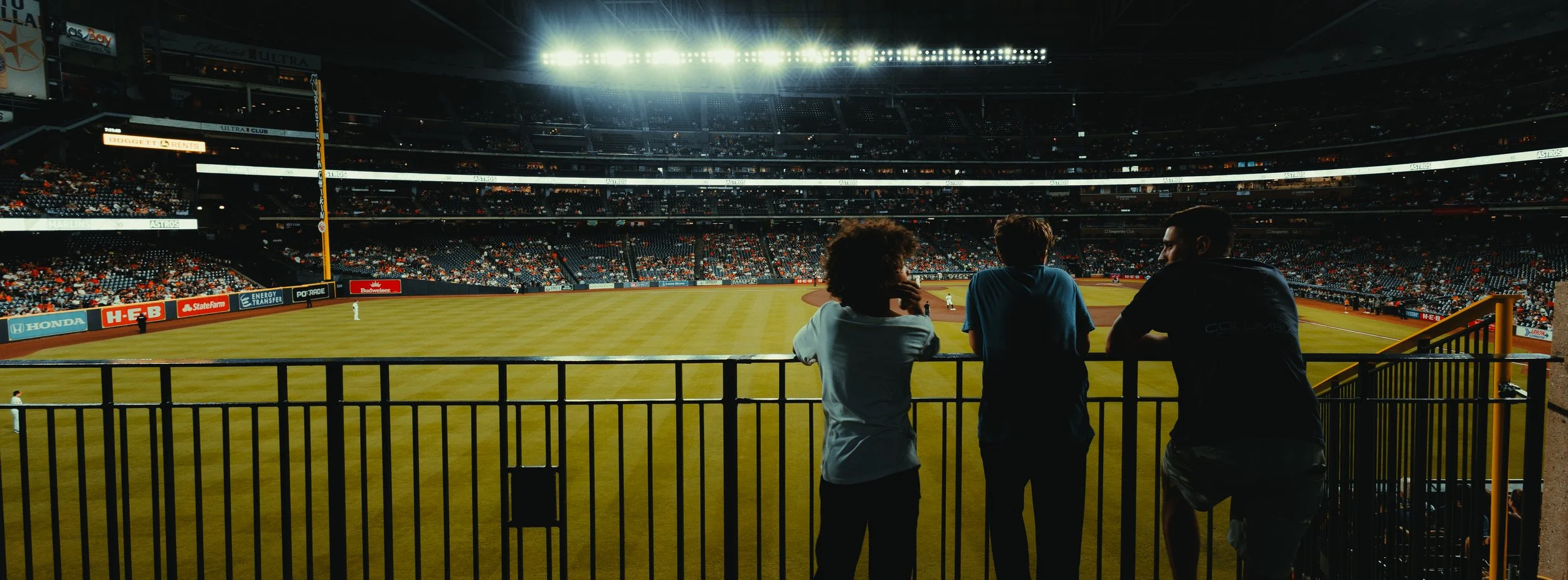 People watching a baseball game at a stadium from a viewing area, illuminated lights overhead and empty seats in the background.