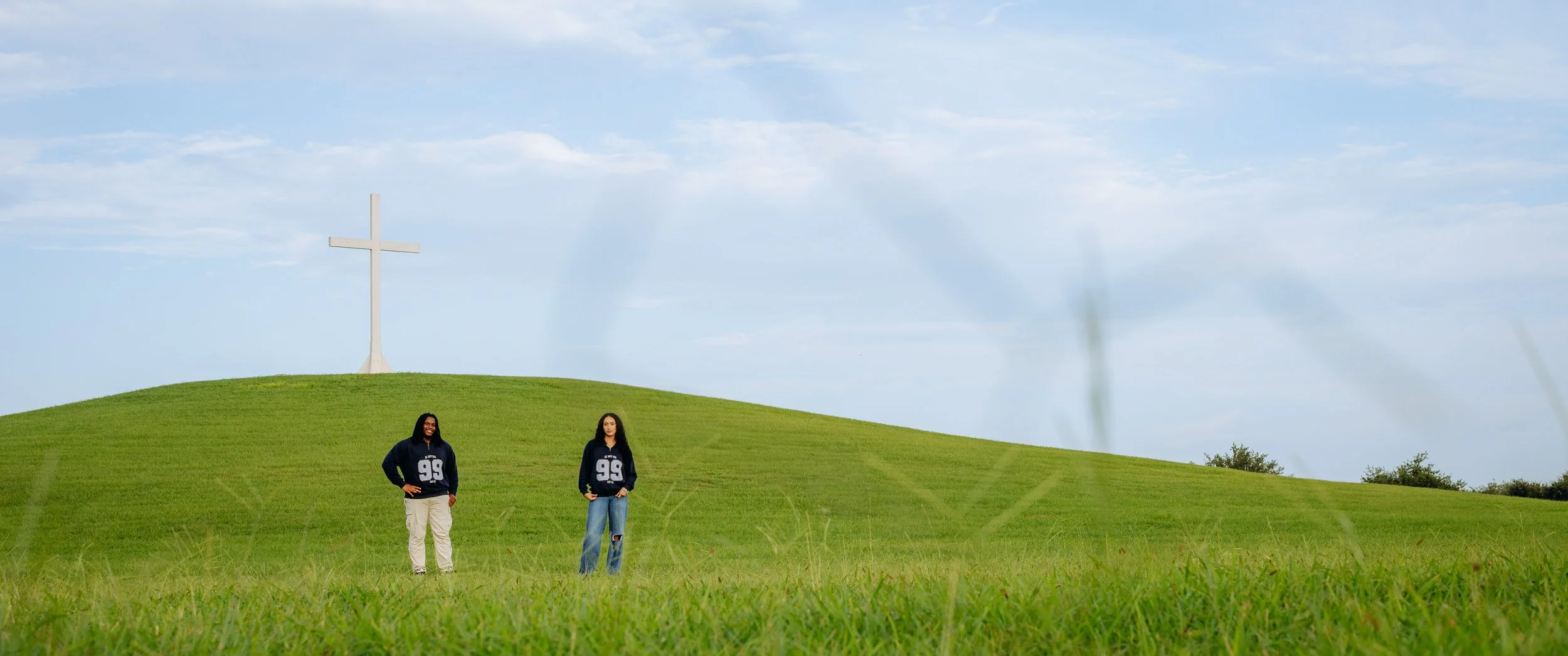 Two people standing on a grassy hill with a large white cross in the background, under a blue sky with clouds.
