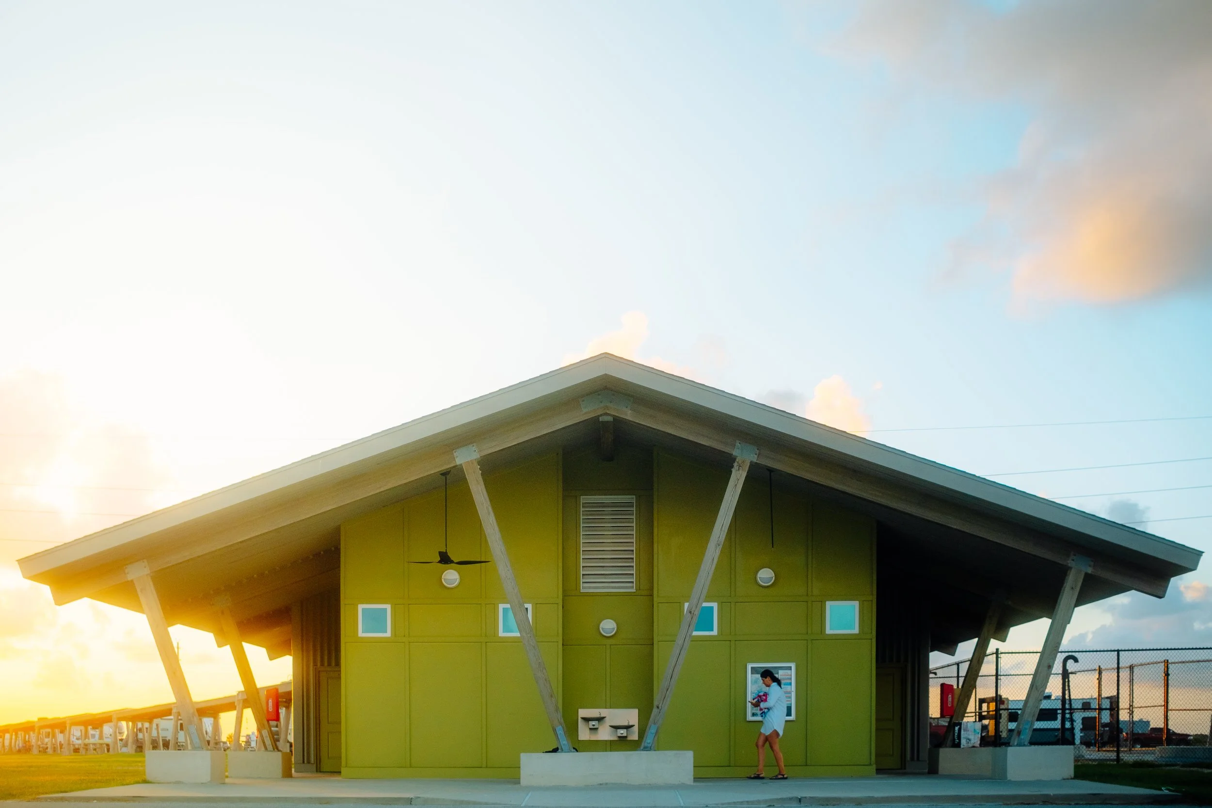 A green public restroom building with a pitched roof, support beams, and small windows, located in a park at sunset, with a person holding flowers standing outside.