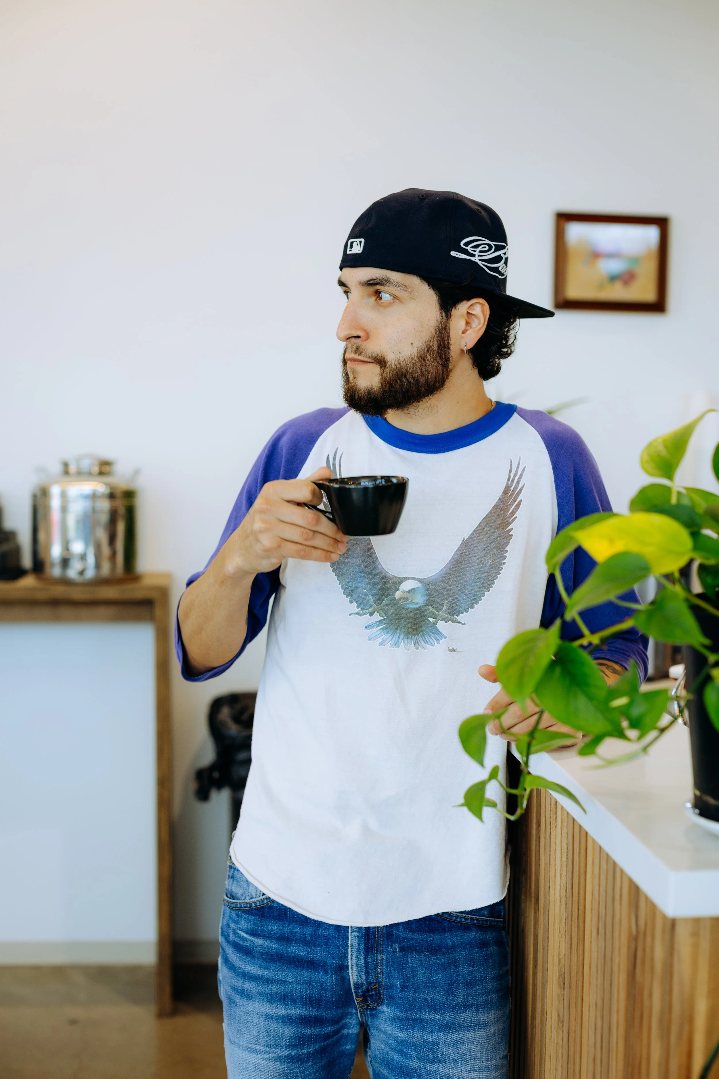 A man with a beard wearing a black baseball cap backwards, a white t-shirt with purple sleeves and an eagle graphic, and blue jeans, standing indoors near a counter with a plant, holding a black cup and looking to the side.