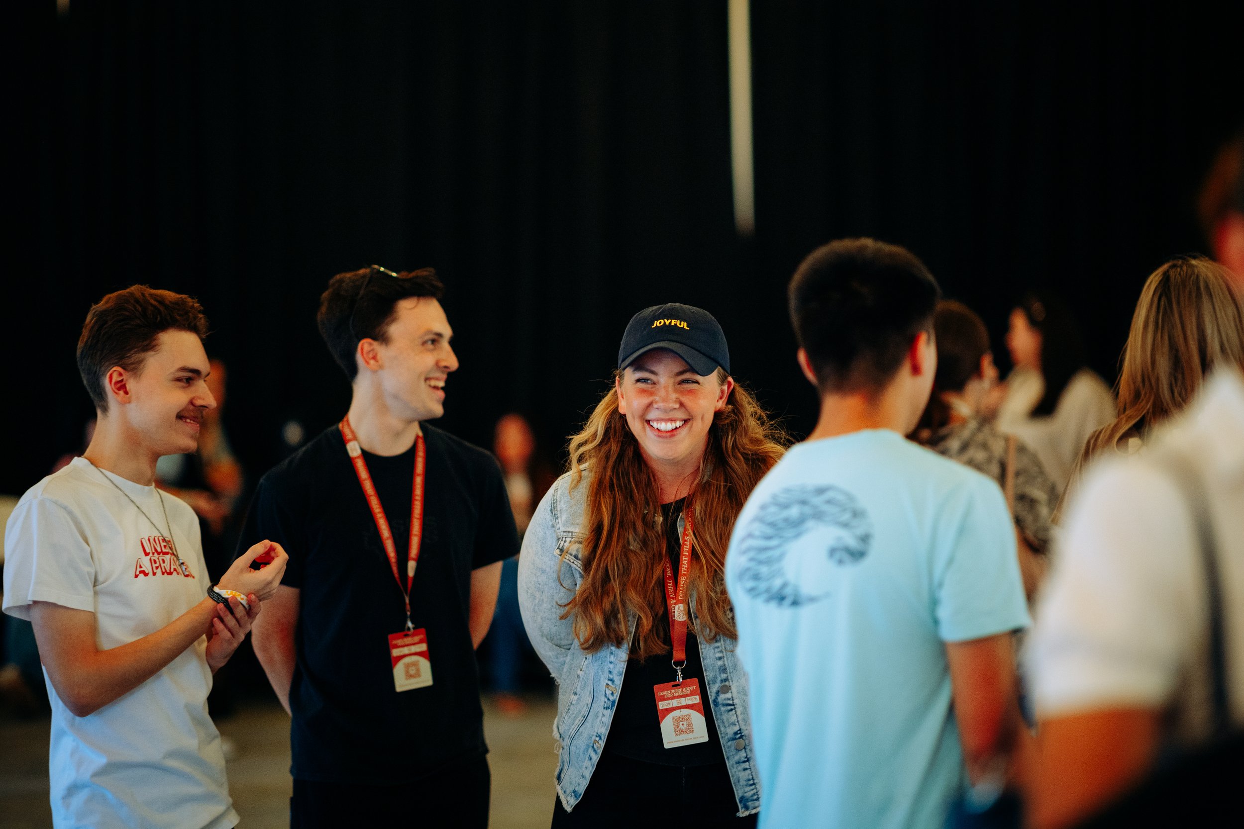Group of young people smiling and conversing at an indoor event, some wearing conference badges and lanyards.