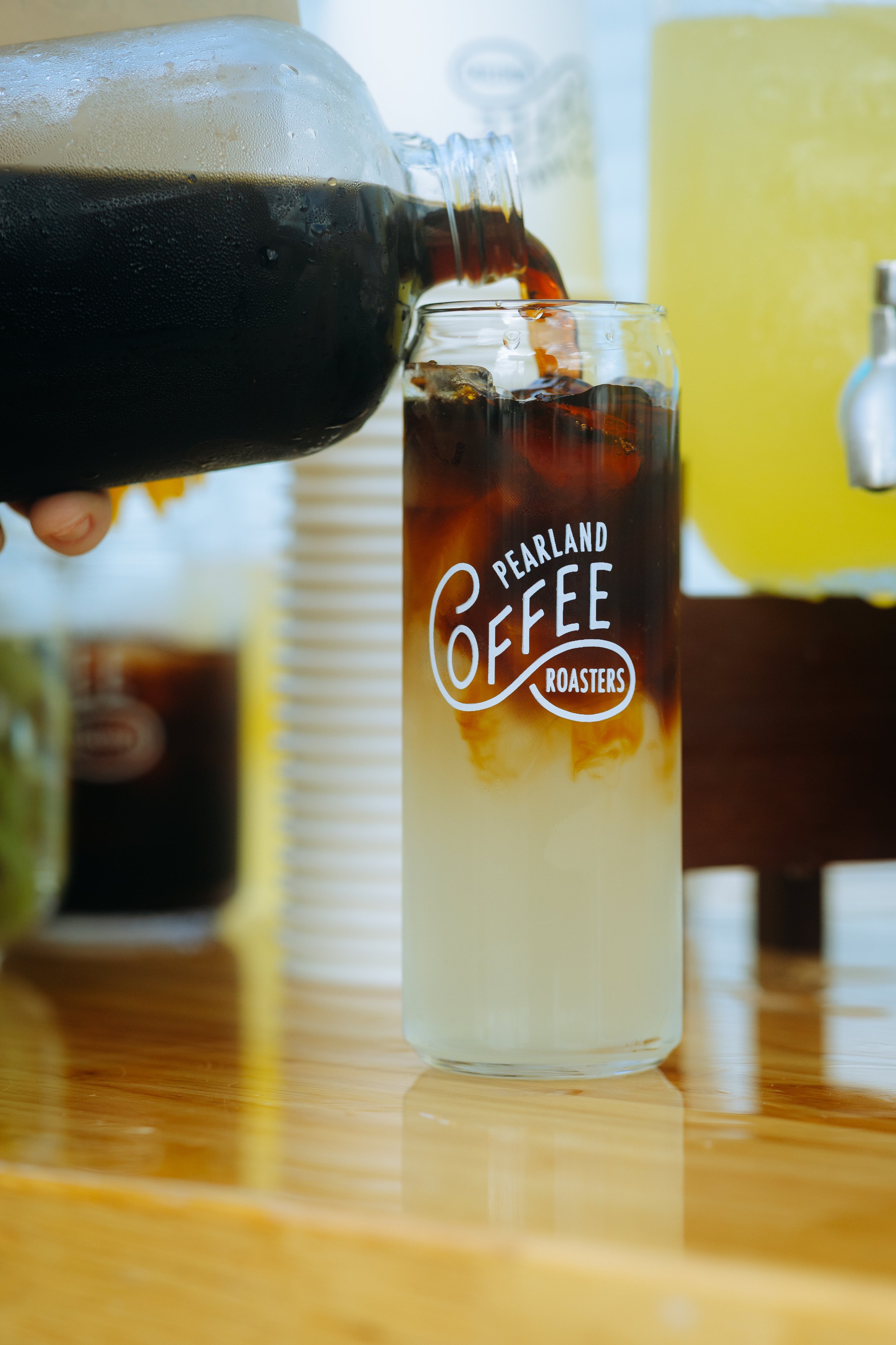 A glass of iced coffee with cream being filled from a coffee pot, labeled 'Pearland Coffee Roasters' on a wooden table.