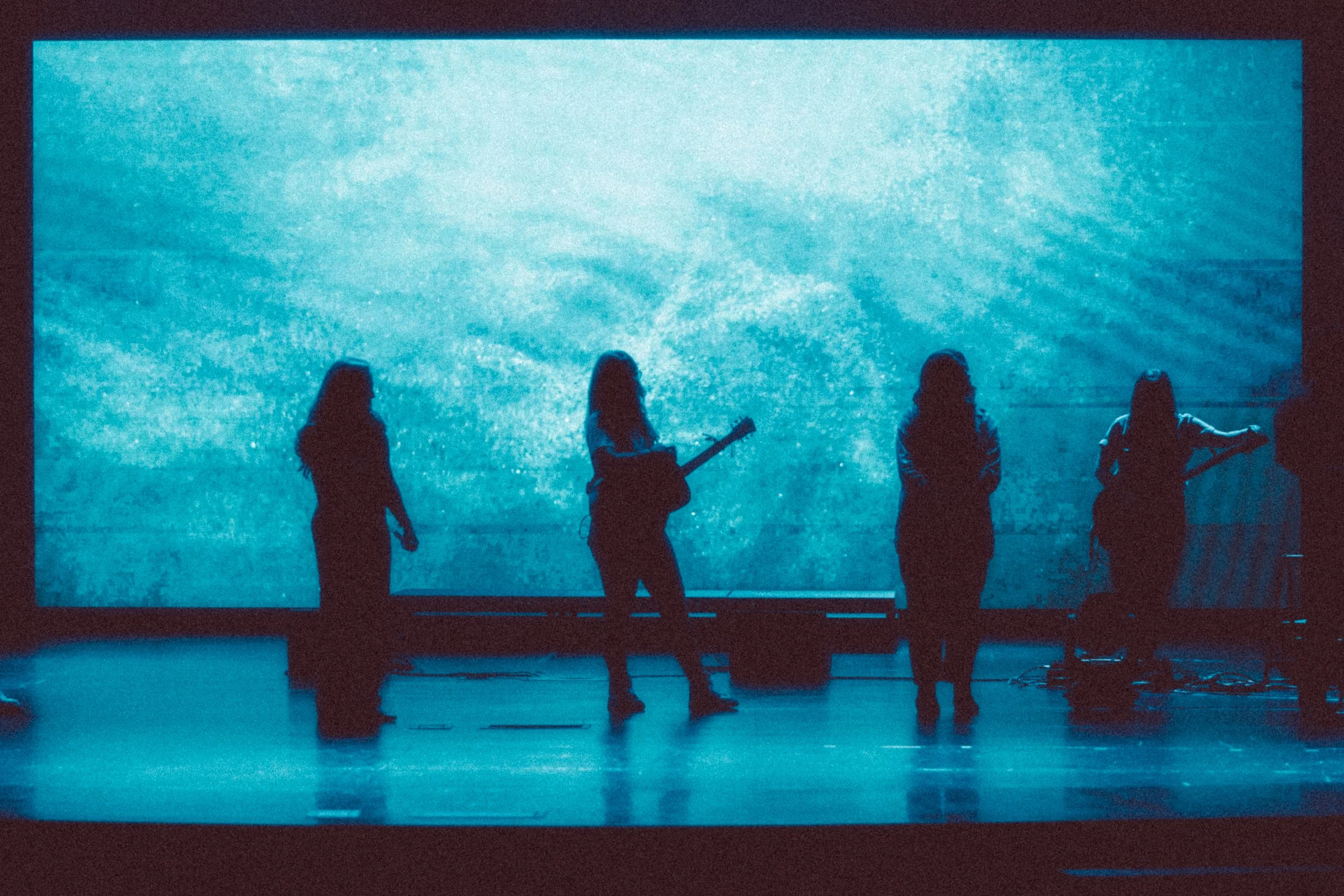 Four women silhouetted against a large blue-screen with a water or ocean texture, two of them holding guitars.