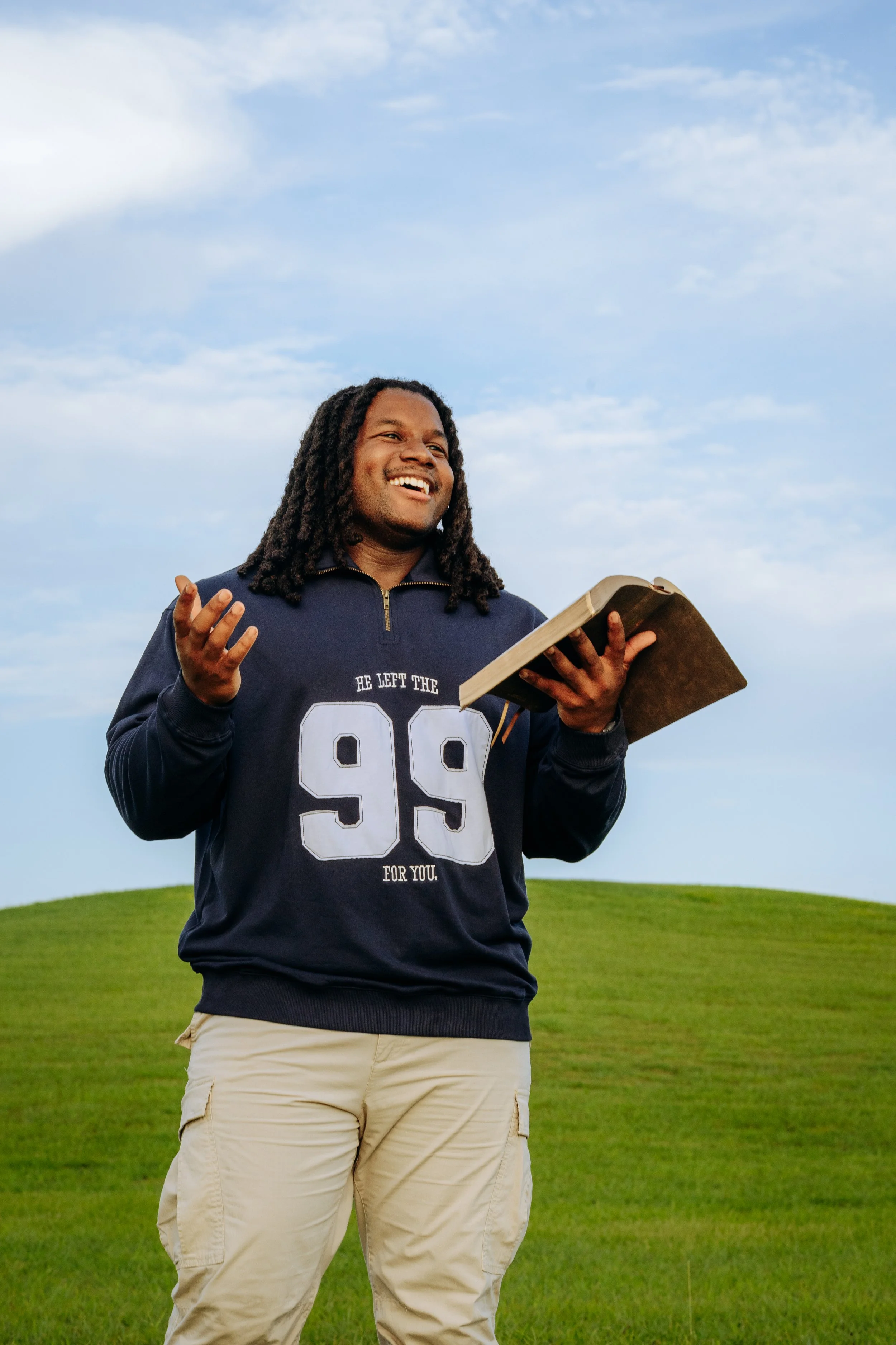 A smiling man with dreadlocks wearing a navy blue sweatshirt and beige cargo pants stands outdoors on a grassy hill, holding an open book or journal, with a blue sky and scattered clouds in the background.