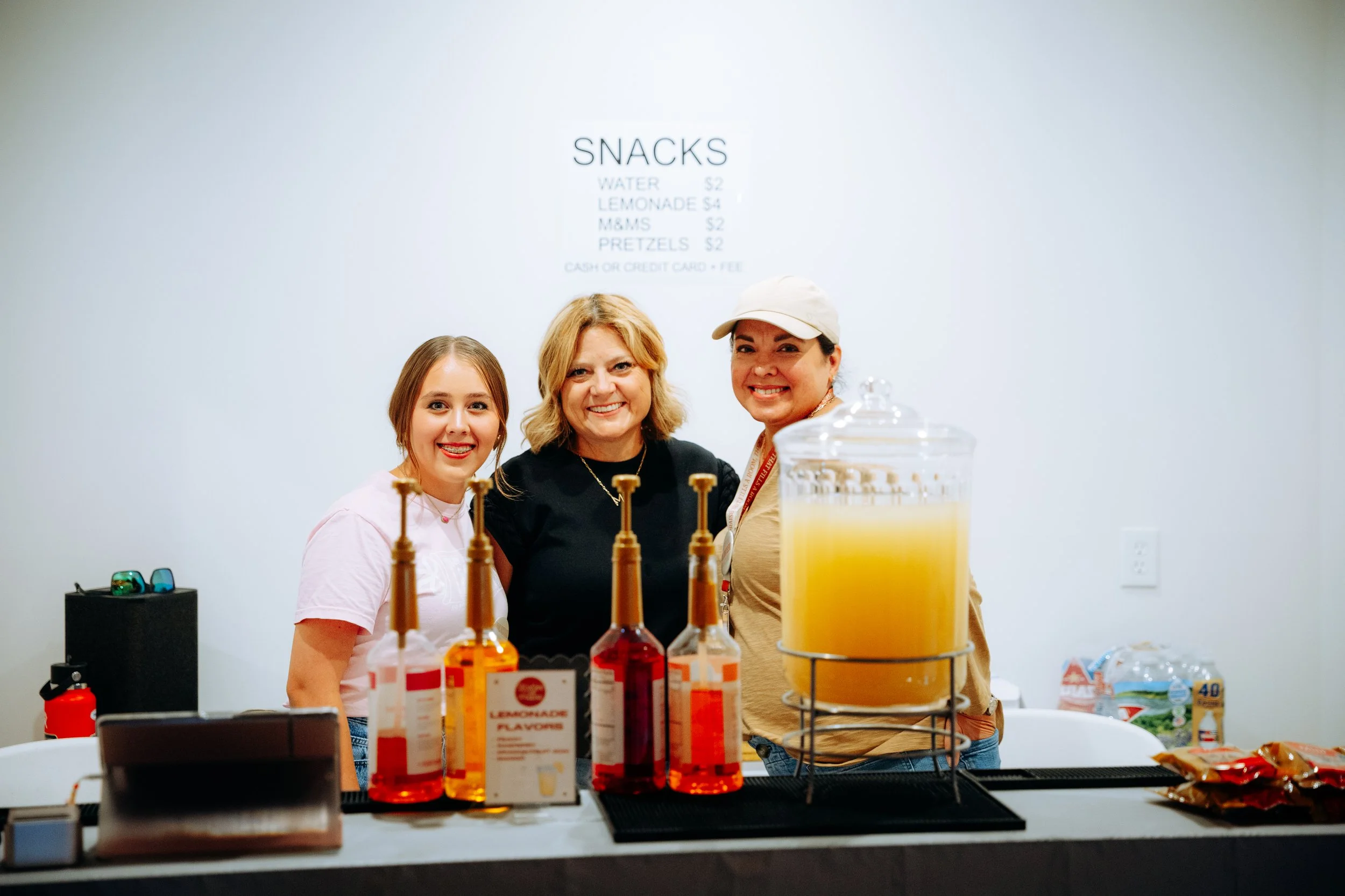 Three women behind a counter with condiments, juice, and snacks in a room with a snack menu on the wall.