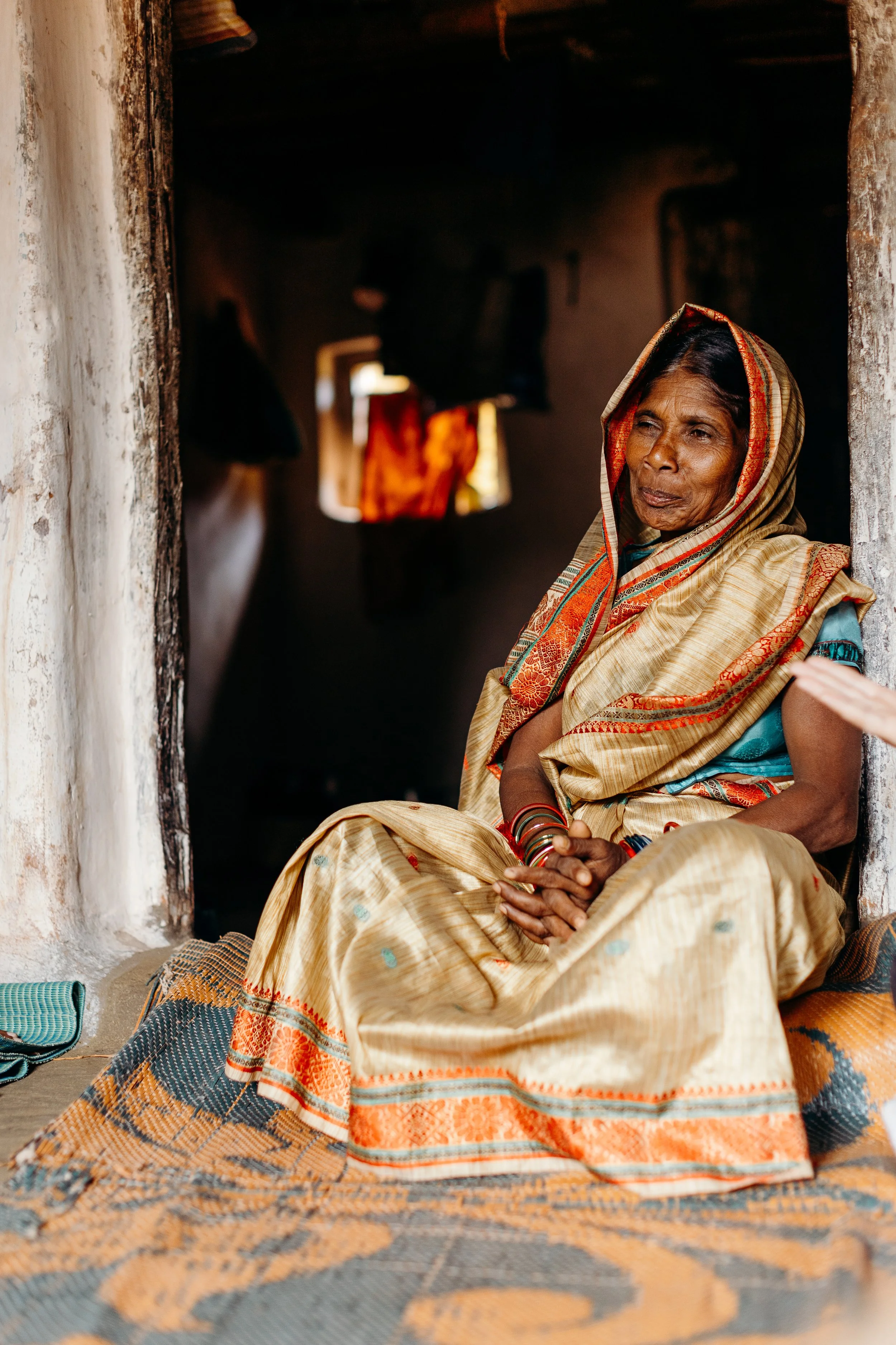 An older woman sitting on the floor in a doorway, wearing traditional Indian attire and colorful bangles, with a dark interior behind her.