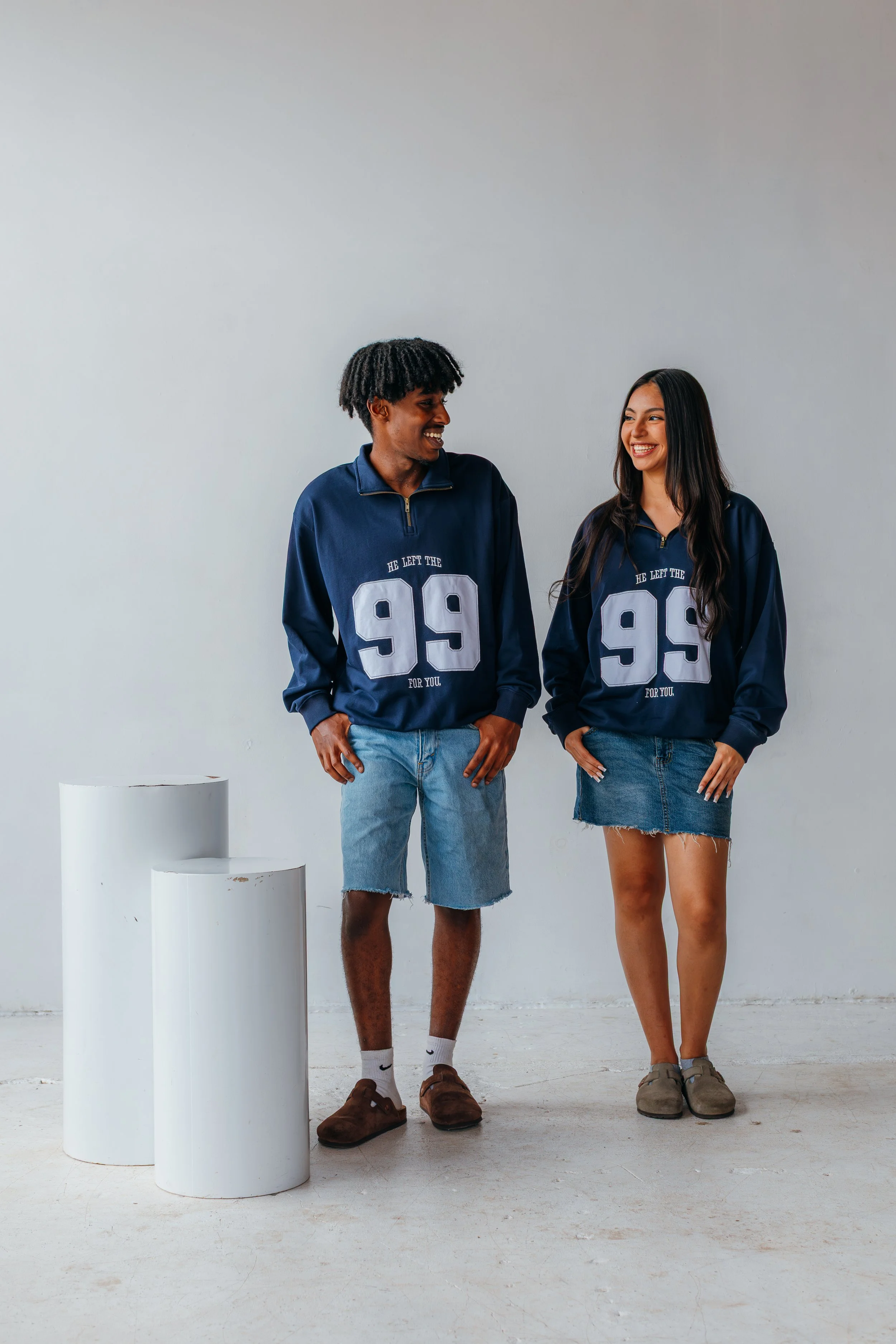 Two young adults, a man and a woman, standing and smiling at each other in a minimalistic, white studio setting. They are wearing matching navy blue sports jackets with the number 99 and the text 'He left the 99 for you.' The man is wearing denim sho