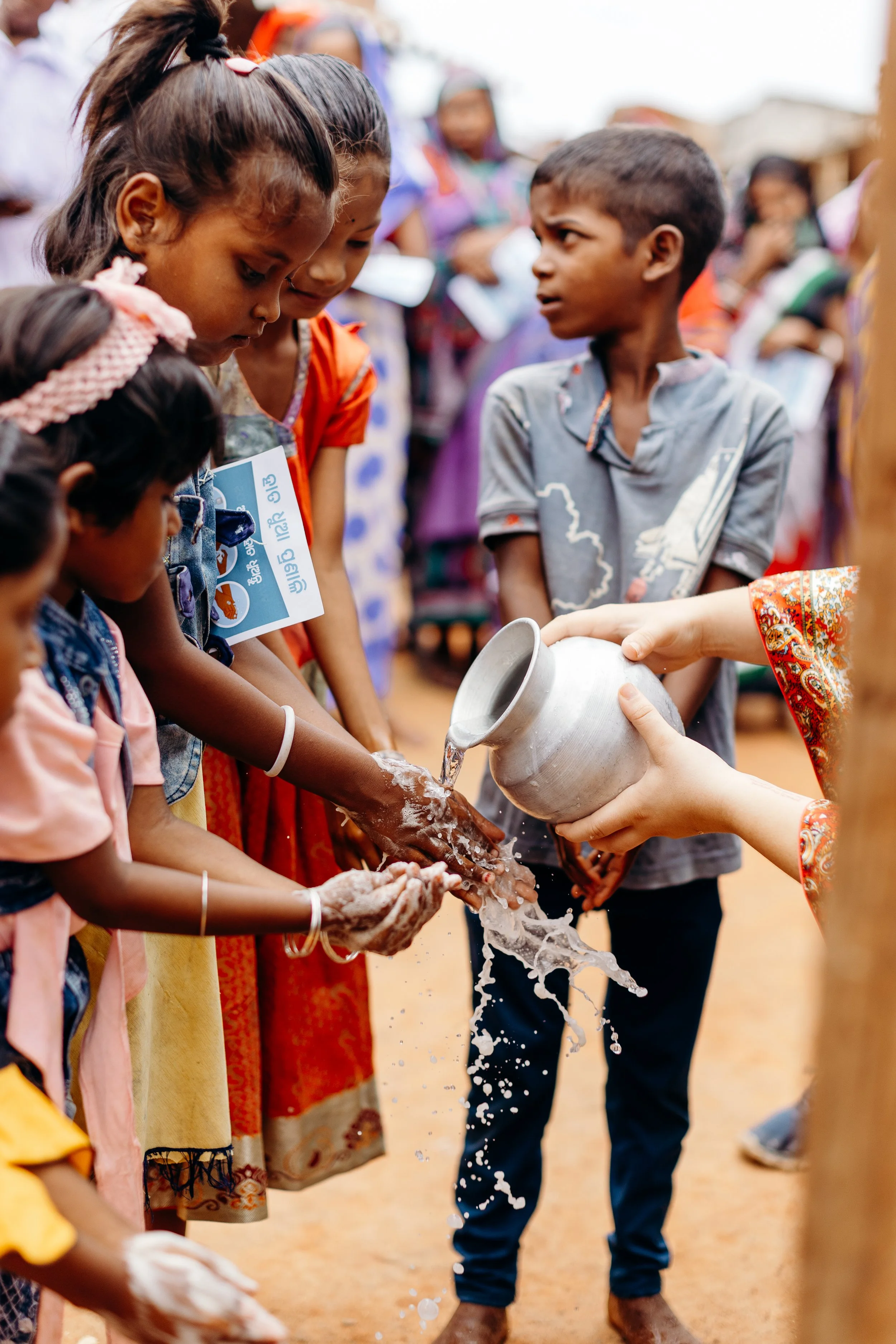 Children washing their hands with water poured from a metal container during an outdoor event.