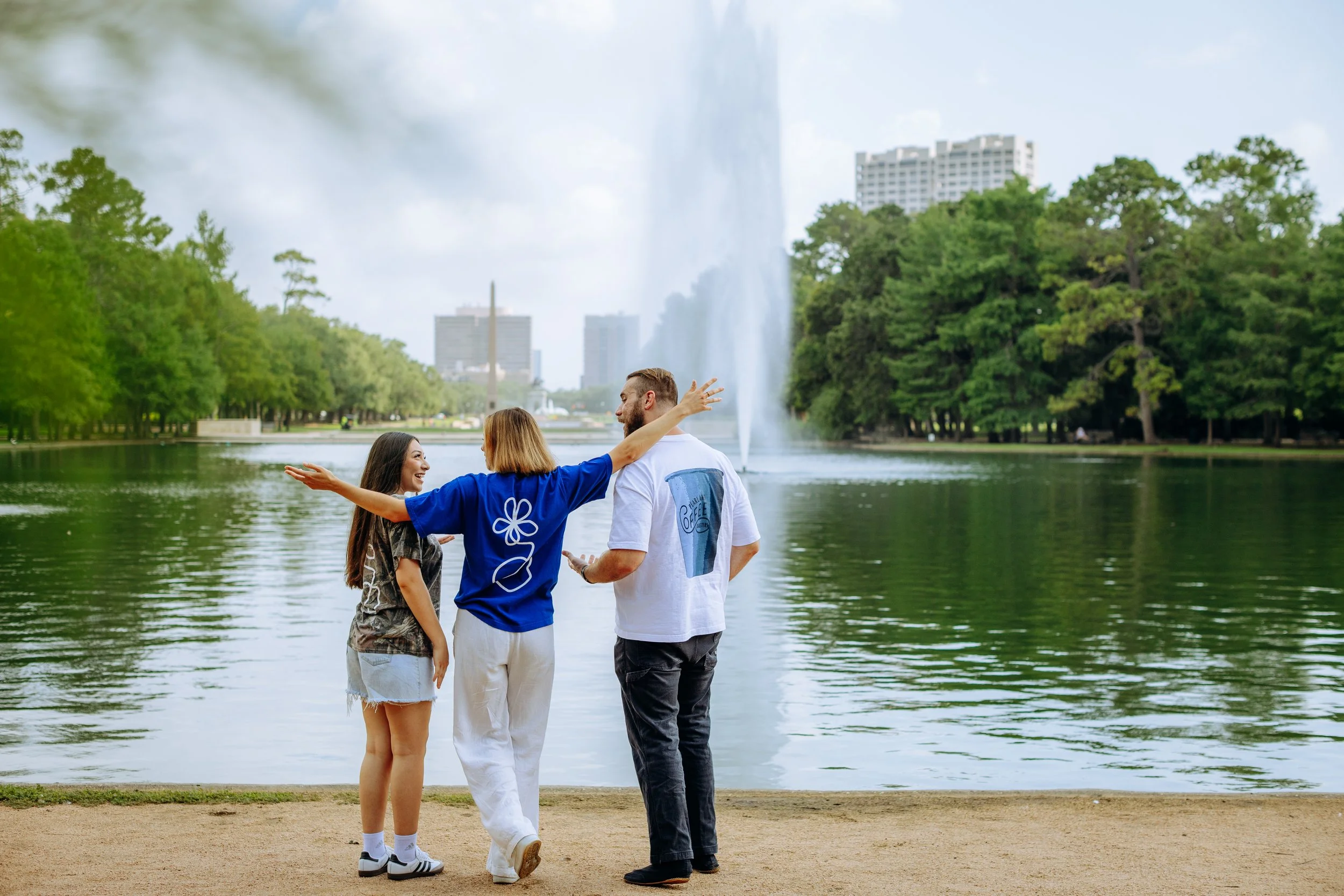 Three people standing near a lake with a fountain, surrounded by trees and tall buildings in the background, smiling and engaging with each other.