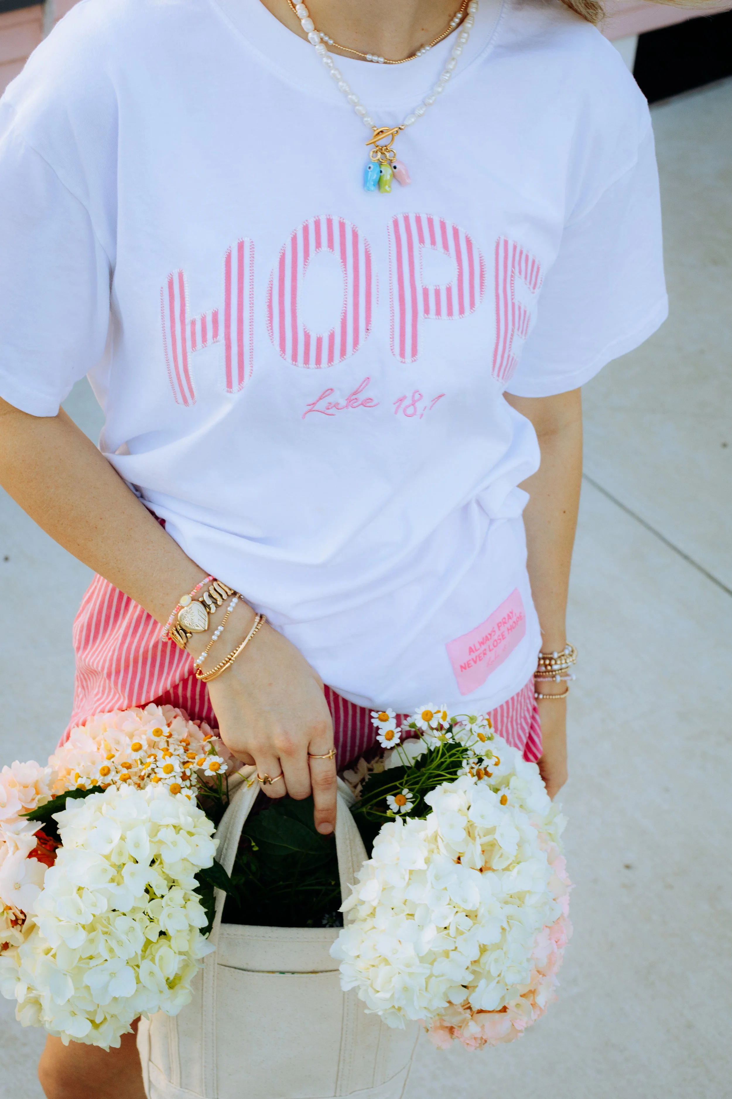 Person wearing a white T-shirt with pink striped letters spelling 'HOPE' and holding a beige bag filled with white and pink flowers.