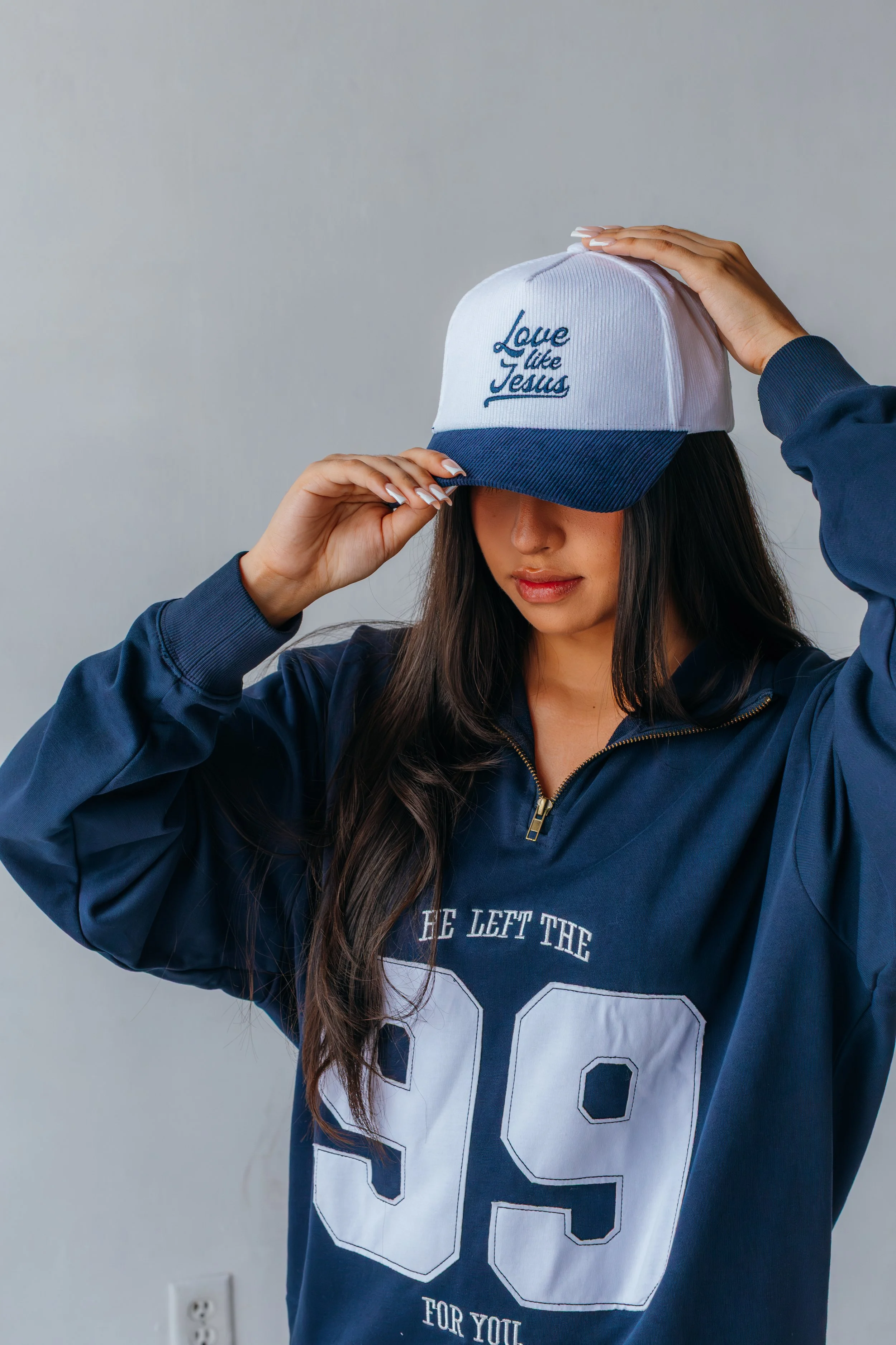A young woman with long dark hair wears a navy blue sports jersey with the number 99 and the text "HE LEFT THE 99 FOR YOU". She is holding a white and blue baseball cap with "Love like Jesus" embroidered on it, tilting it down over her eyes, against 