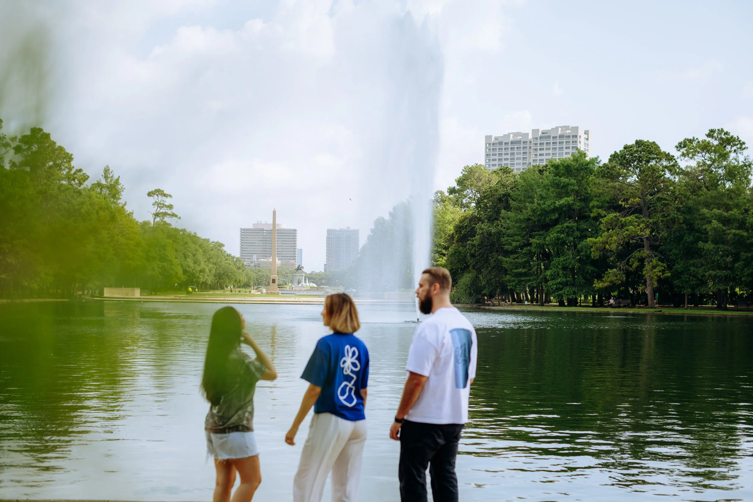 Three people standing by a lake in a park with trees, a fountain, and tall buildings in the background.