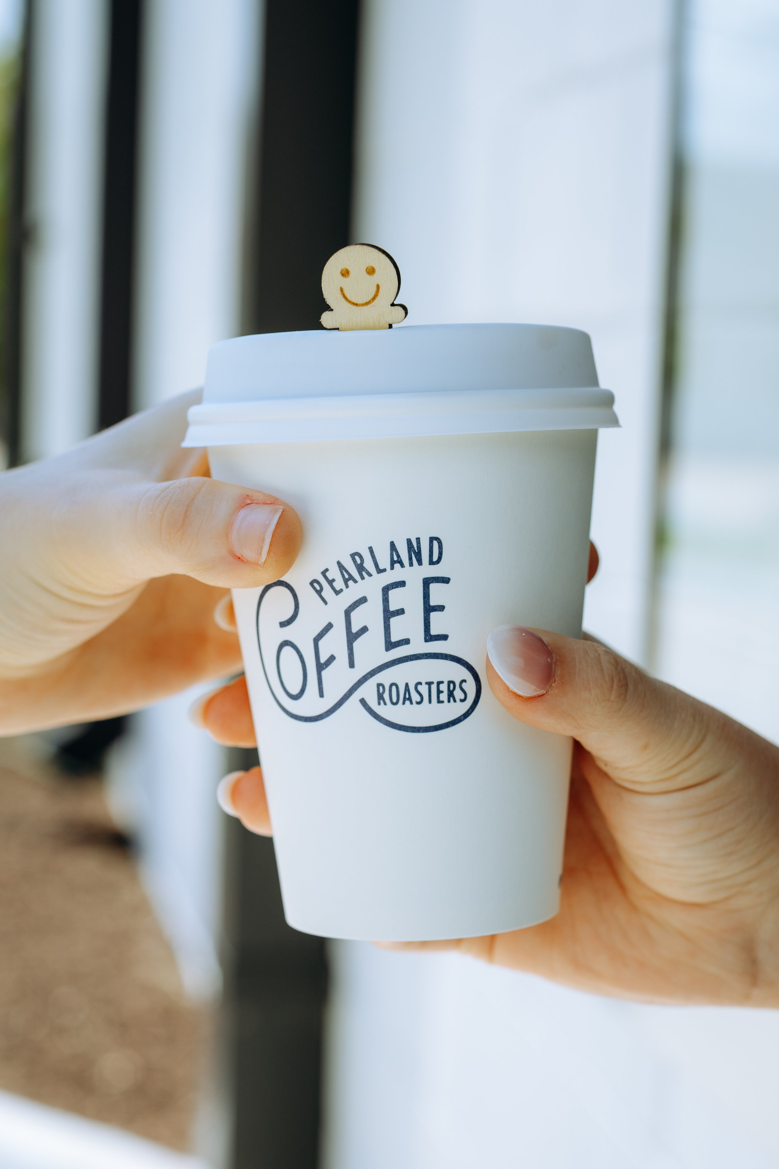 Person holding a white paper coffee cup with 'Pearland Coffee Roasters' written on it, and a small wooden smiley face figure on the lid.