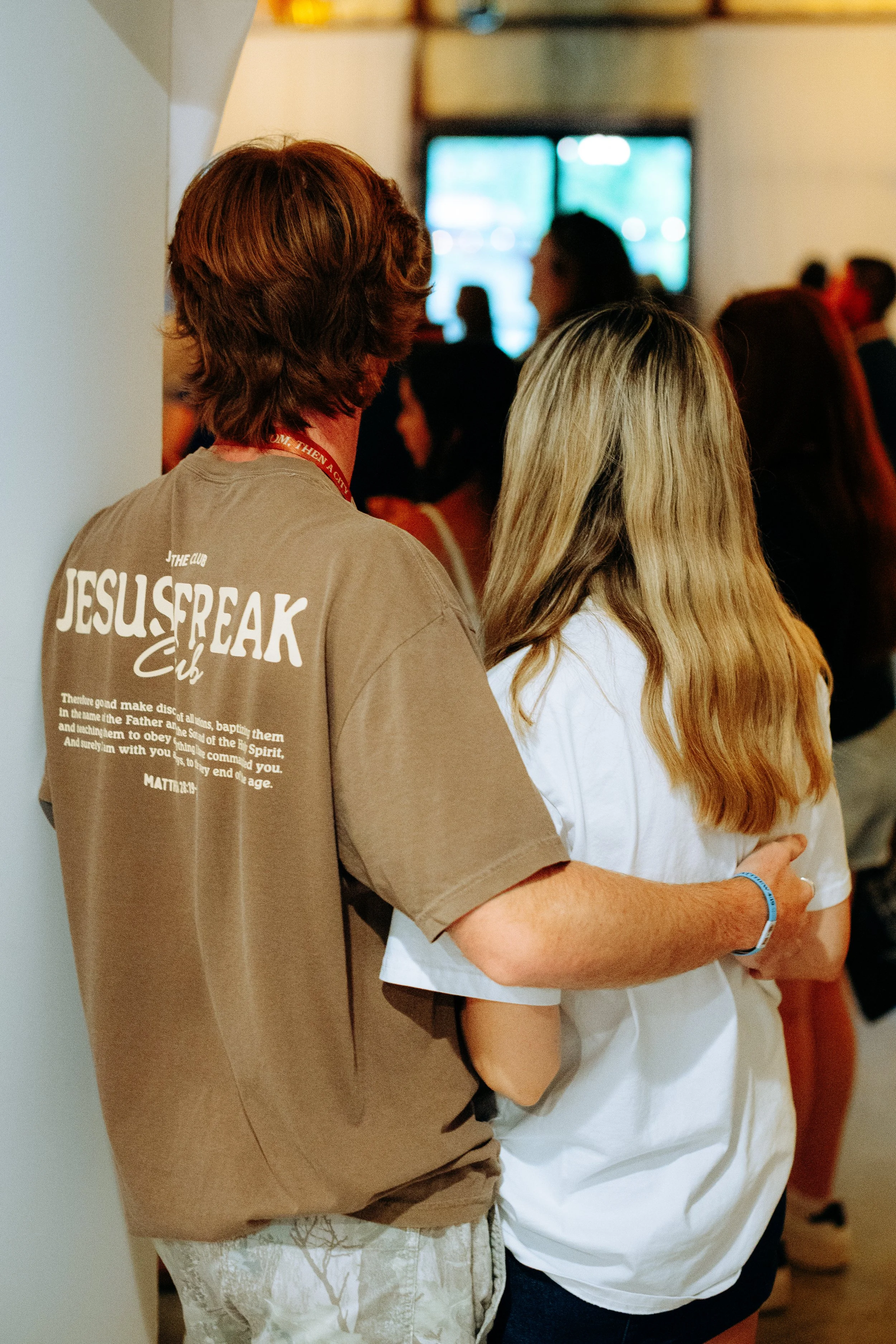 A young man and woman standing close together in a crowd, with the man’s arm around the woman’s waist, at an indoor event. The man is wearing a brown T-shirt with the words "JESUS FREAK" on the back, and the woman has long blonde hair and a white shi