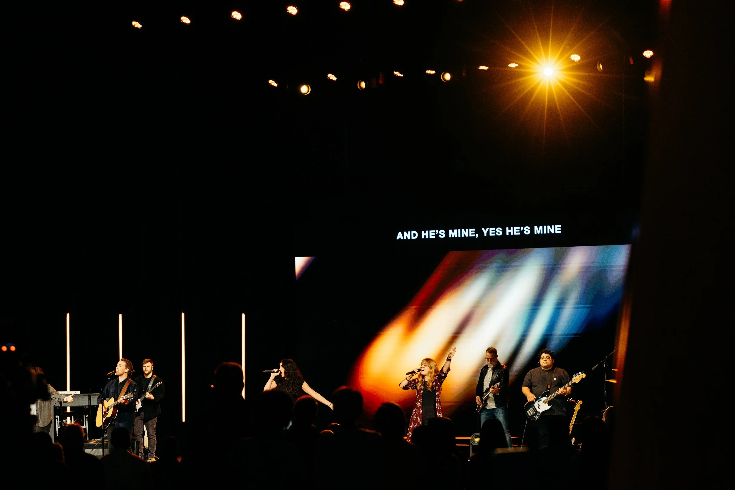 A band performing on stage with five members, including two singers, with a large screen behind them displaying the text "And he's mine, yes he's mine." The stage is lit with yellowish spotlights, and an audience is watching the performance.