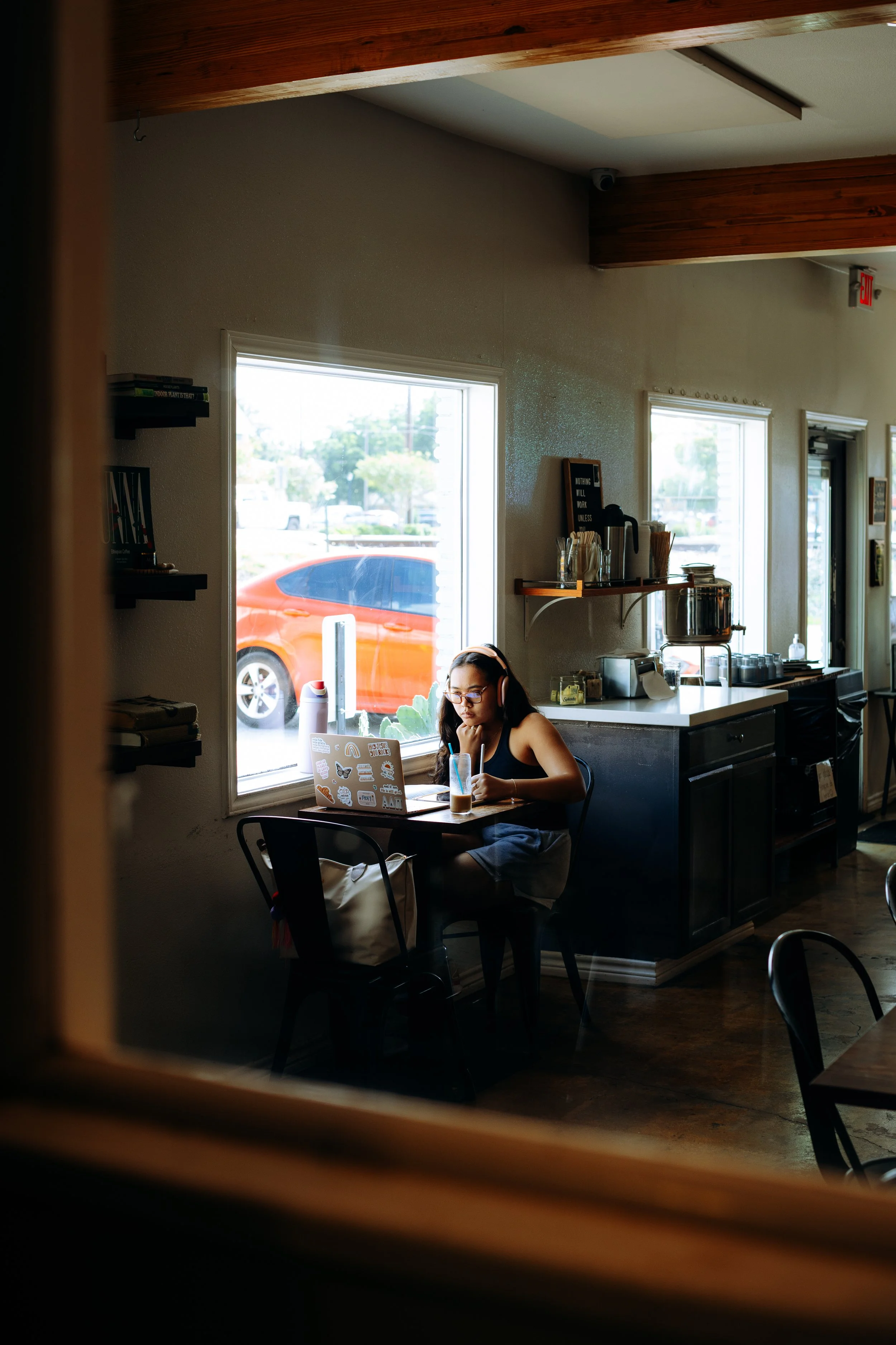 Young woman sitting alone at a small table in a cozy coffee shop, working on a laptop with stickers, wearing headphones, with a drink in front of her, near a window with sunlight.