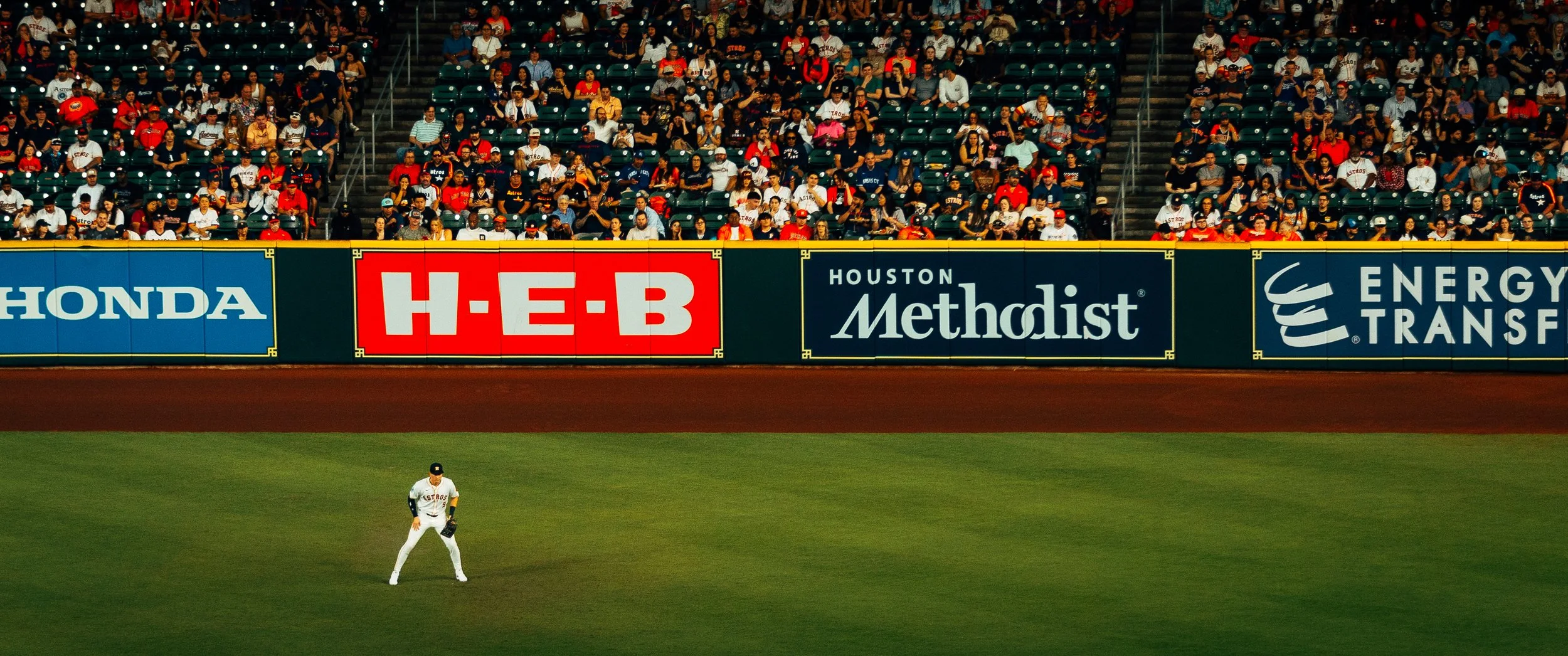 A baseball game in progress with a player on the field and a large crowd in the stands. Advertising banners are displayed on the outfield wall, including H-E-B, Houston Methodist, and Energy Transfer.