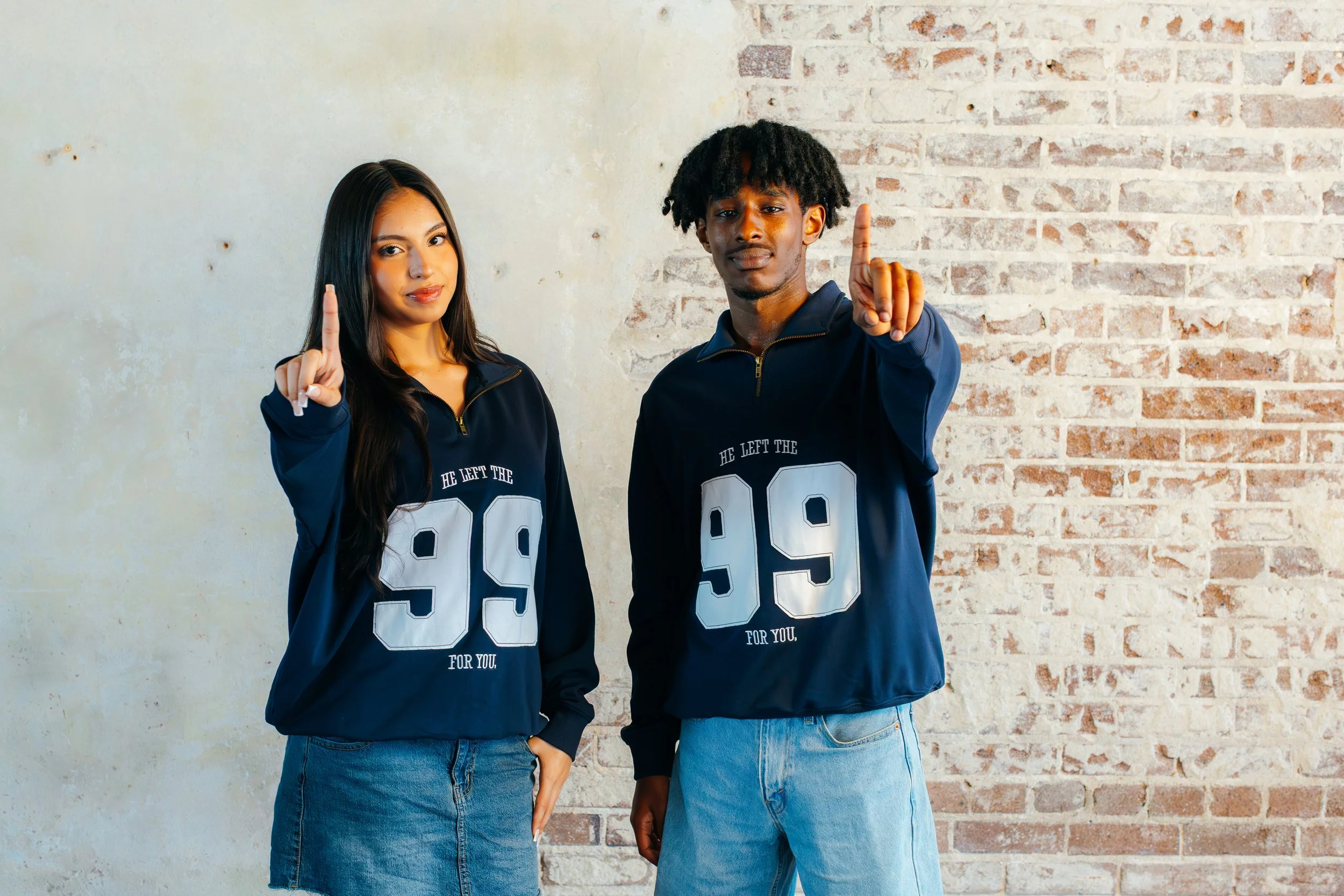 A young woman and young man standing side by side against a brick wall, each wearing a navy blue jacket with the number 99 printed on it, holding up their index fingers.
