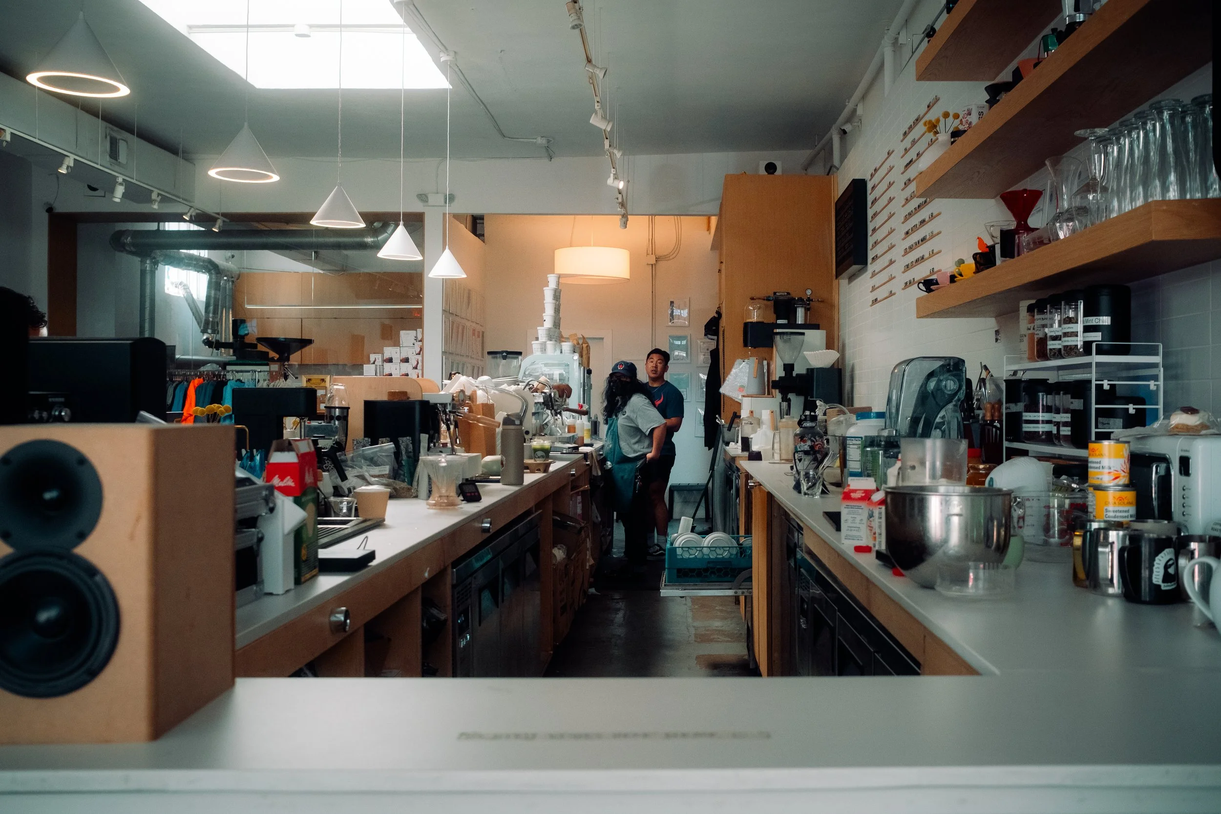Interior of a busy coffee shop with two employees talking near the back, surrounded by coffee brewing equipment, supplies, and kitchen tools.