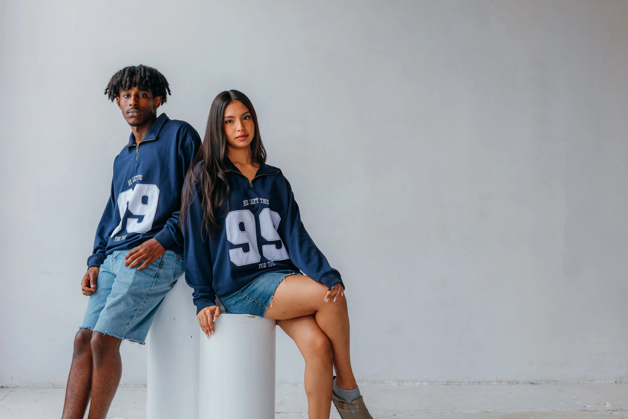 A young man and woman are posing together against a plain light-colored wall. The man is standing with his hand in his pocket, wearing a navy blue sports jacket and denim shorts. The woman is sitting on a white cylinder, wearing a matching navy blue 