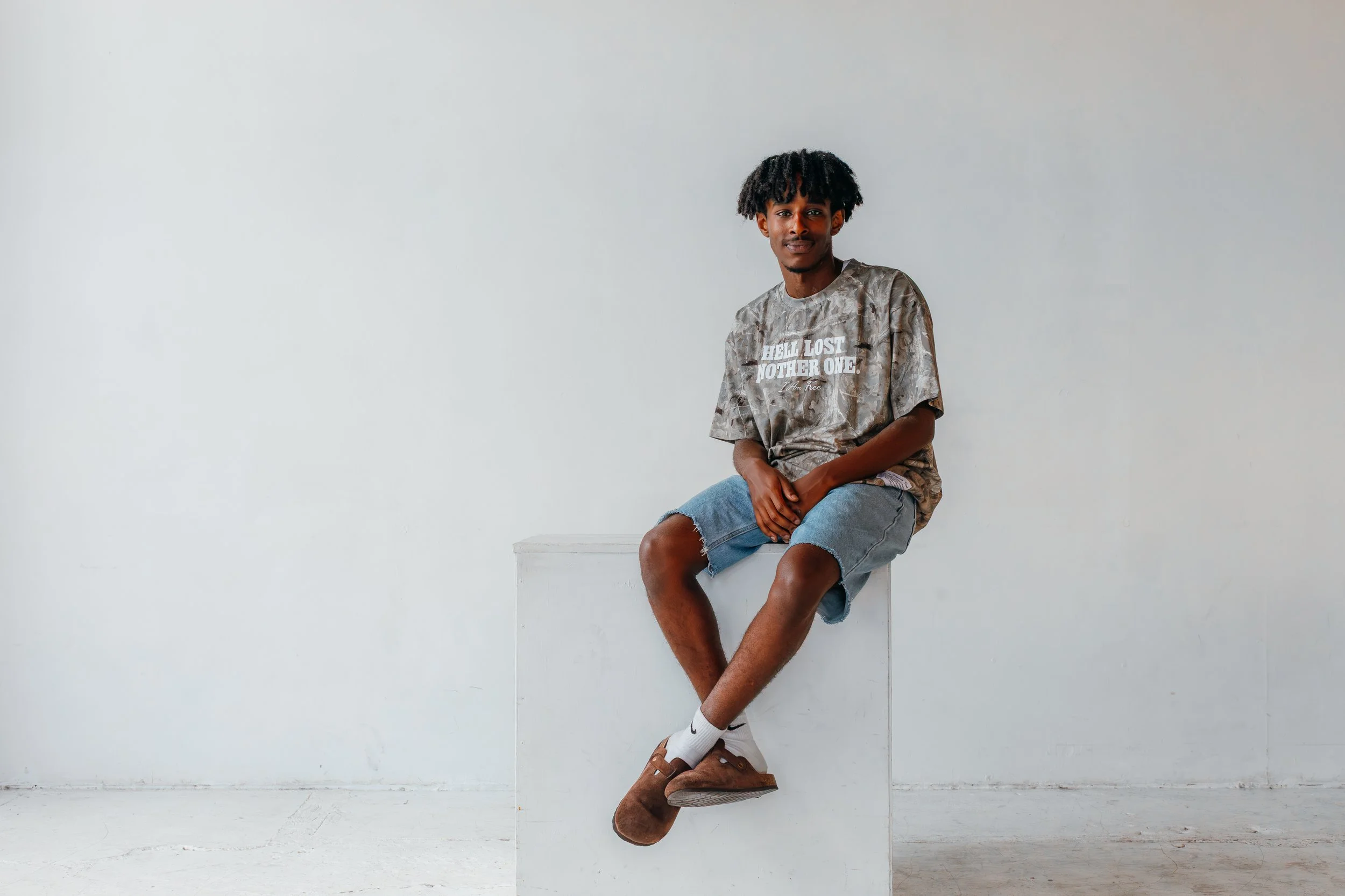 Young man with dark curly hair sitting on a white cube in a minimalist space with white walls, wearing a gray tie-dye t-shirt, denim shorts, white socks, and brown slip-on shoes.