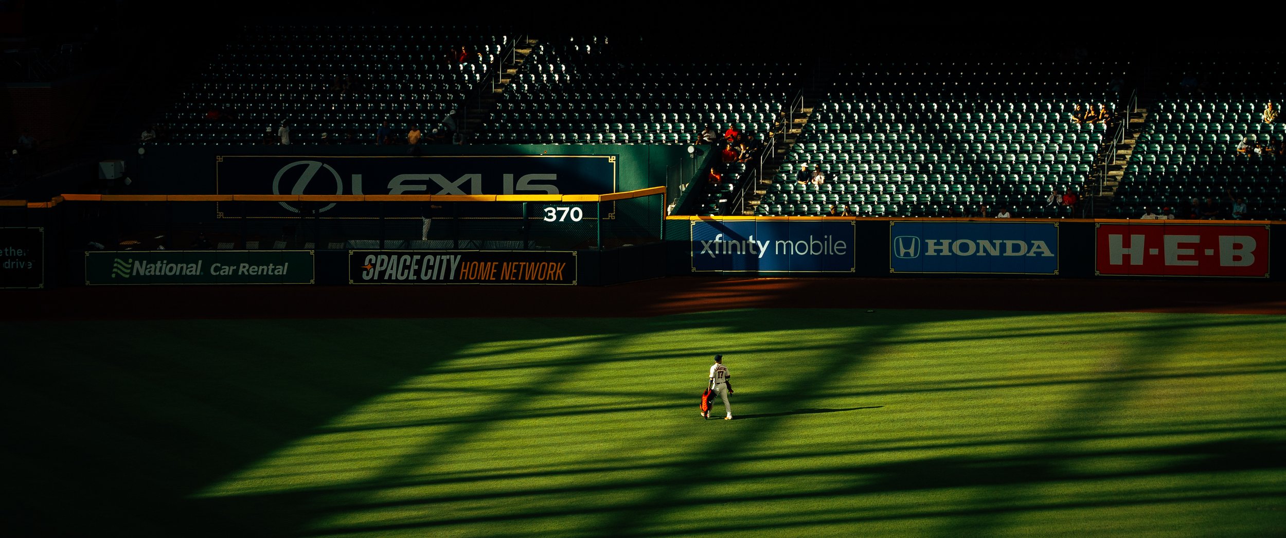 A baseball player walking on the field in the early evening sunlight, with mostly empty stadium seats in the background and advertisements for Lexus, Honda, and other brands on the outfield wall.