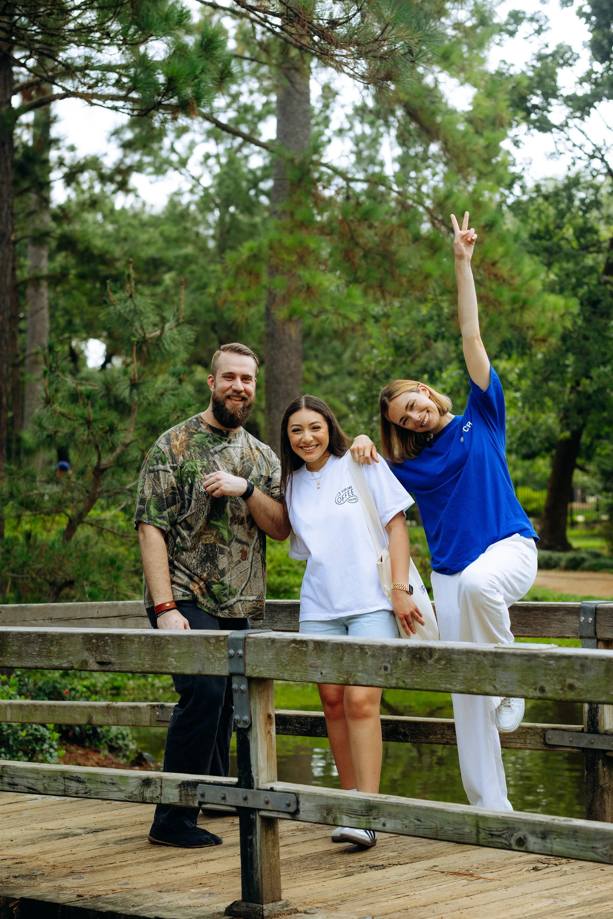 Three friends smiling on a wooden bridge in a park with tall trees, one woman making a peace sign, all casually dressed.