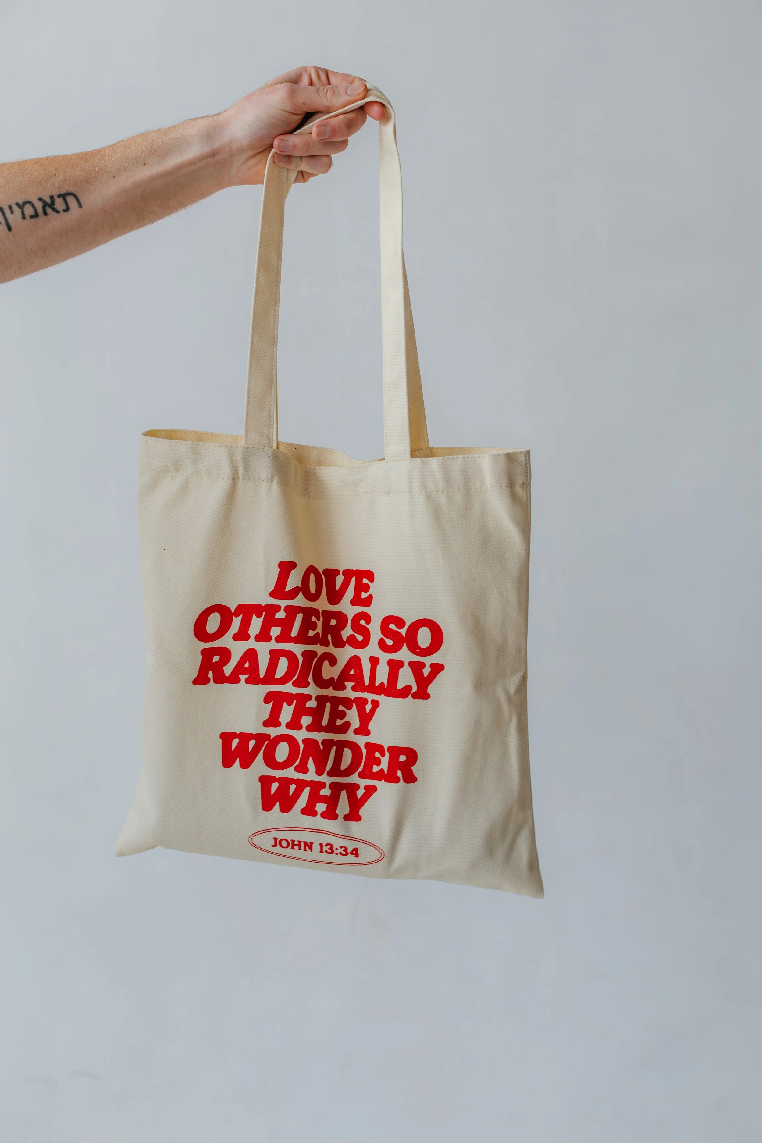 A beige tote bag with red text reading "LOVE OTHERS SO RADICALLY THEY WONDER WHY" and a Bible verse reference "JOHN 13:34" below it. The tote is being held by a hand with a tattoo on the forearm.