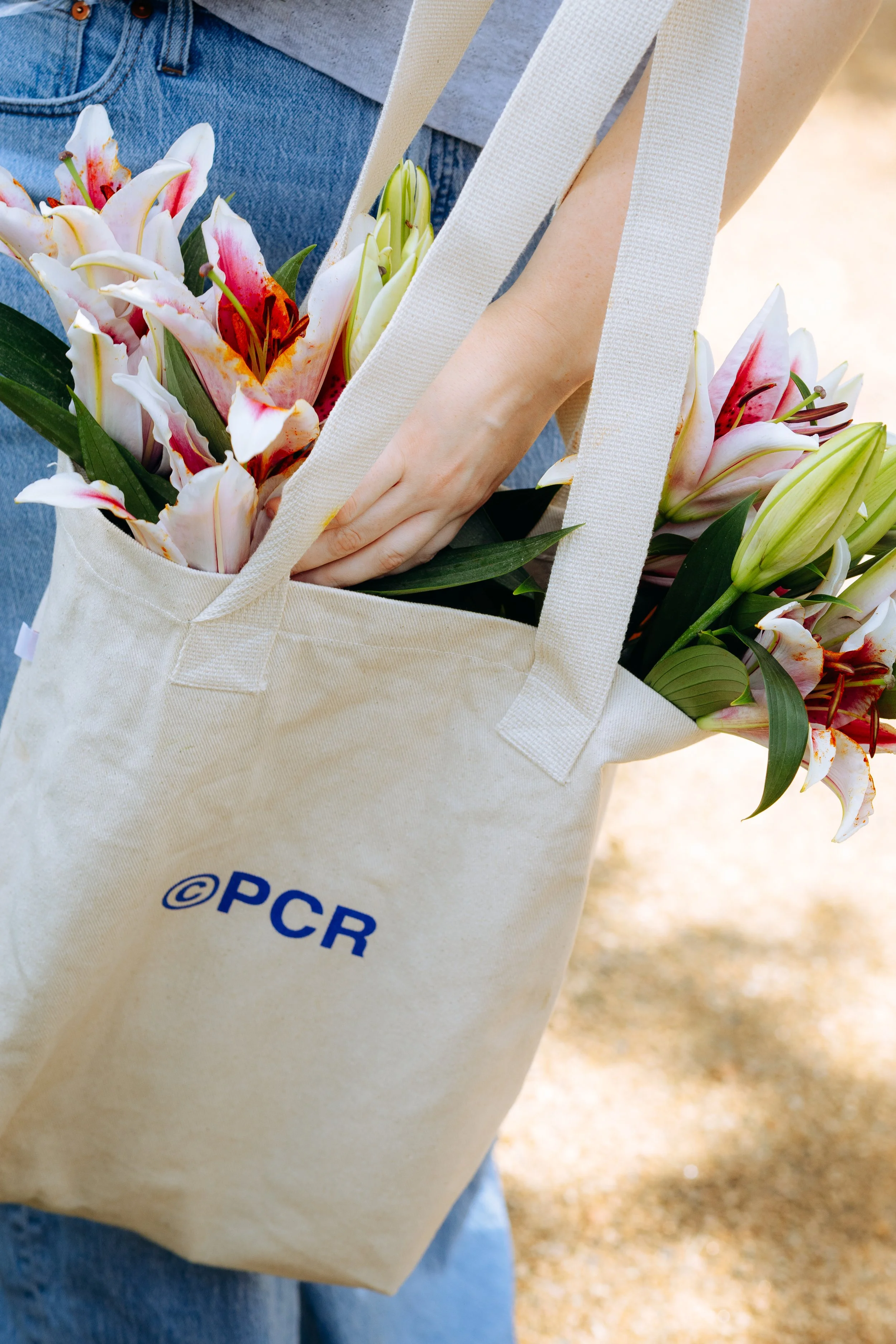 A beige tote bag with blue text “©PCR” holds a bouquet of pink and white lilies, with a person wearing blue jeans standing outdoors.