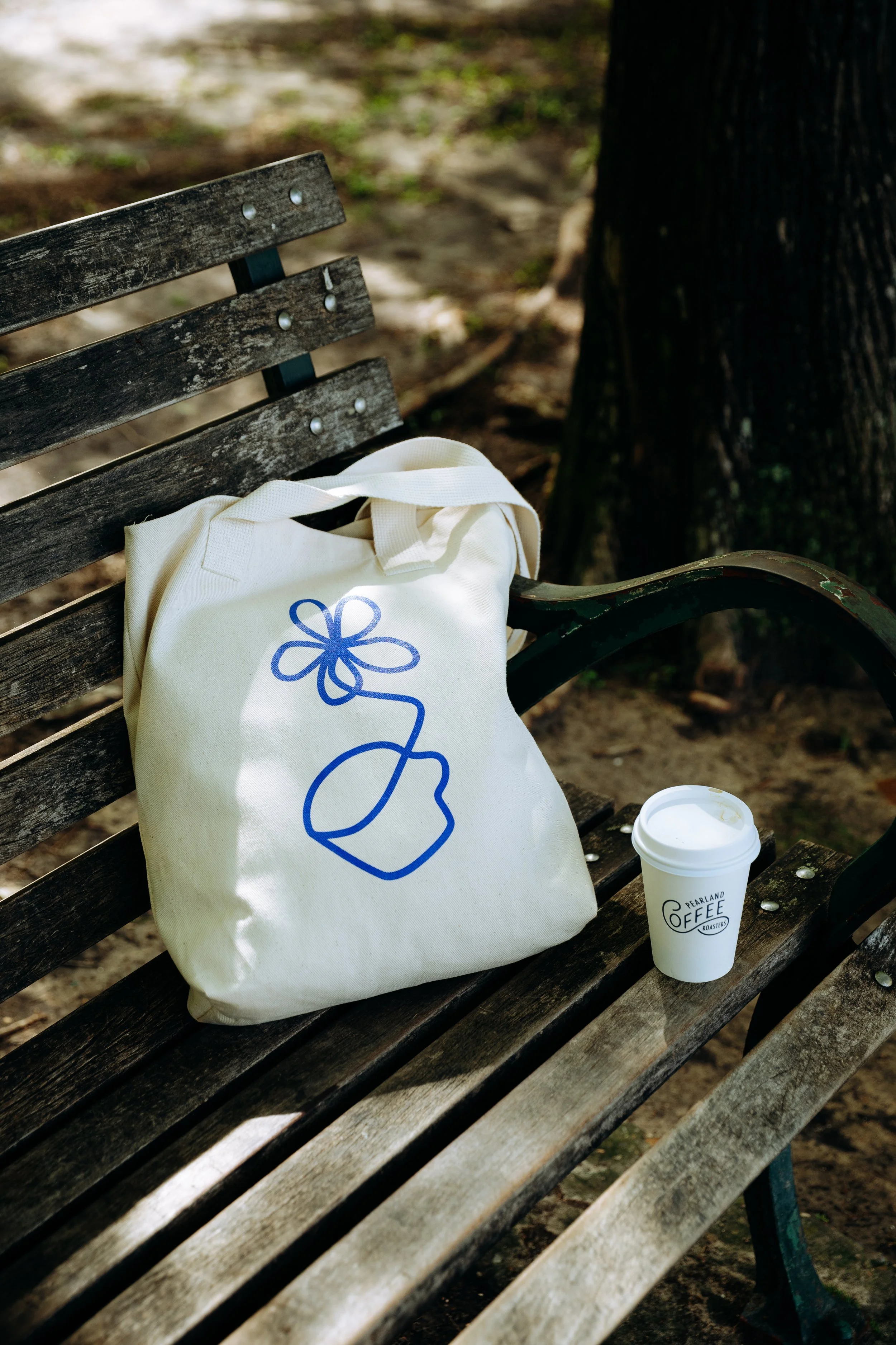 A beige tote bag with blue line art of a cup and flower design and a white paper coffee cup with a lid on a wooden park bench.