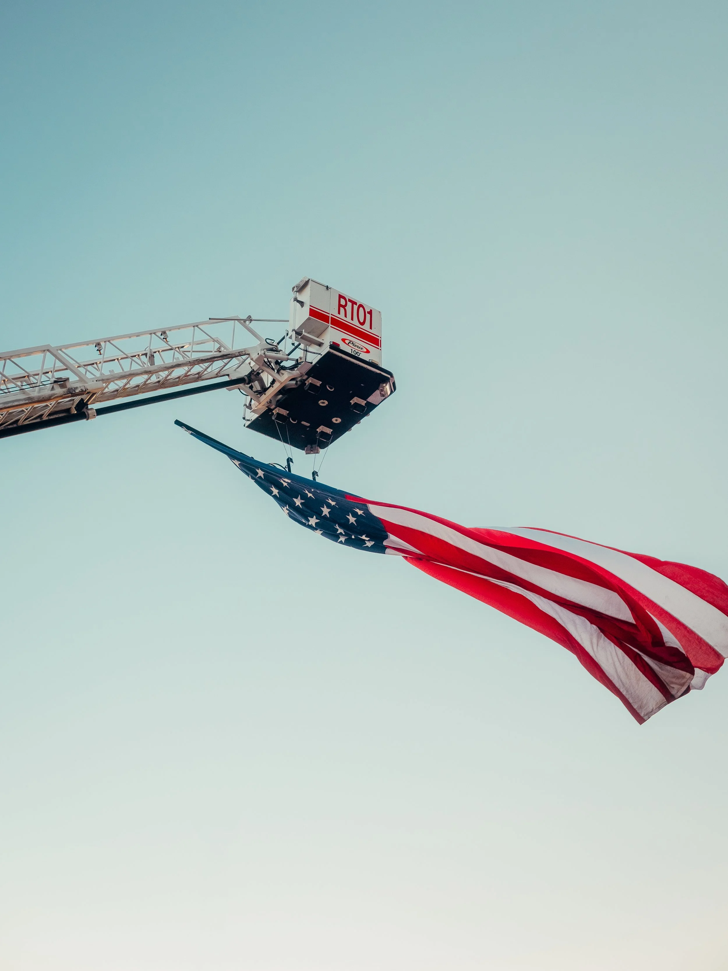 An American flag hanging from a fire truck ladder against a clear sky.