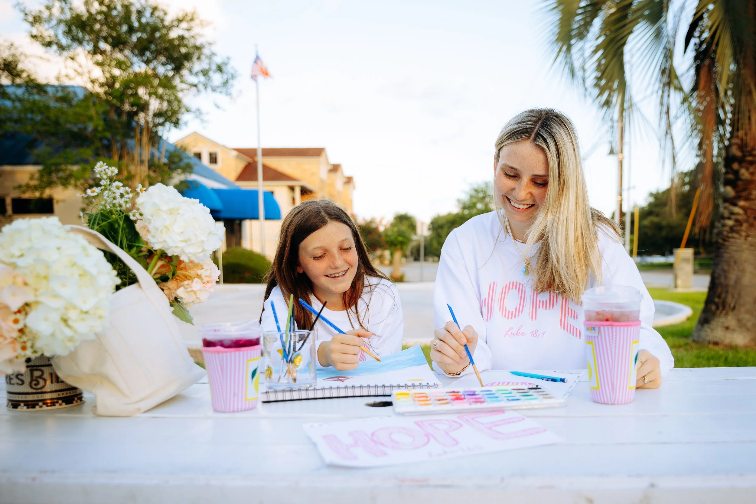 A woman and a girl sitting at a white outdoor table, painting together. The table has flowers, cups with pink stripes, and a watercolor palette. They are smiling and enjoying a bright, sunny day.