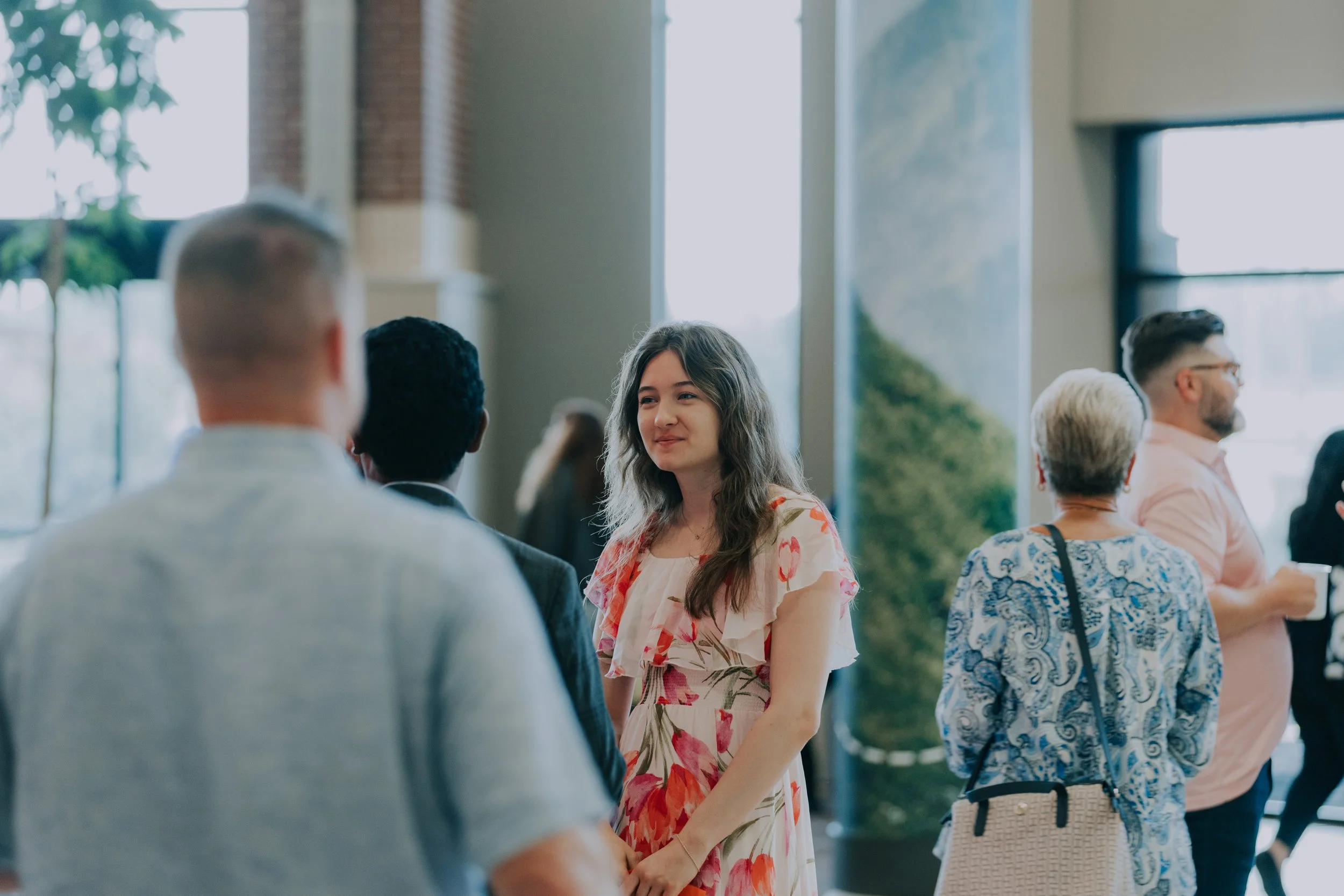Group of people socializing indoors at a bright event, including a woman in a floral dress smiling and talking to people.