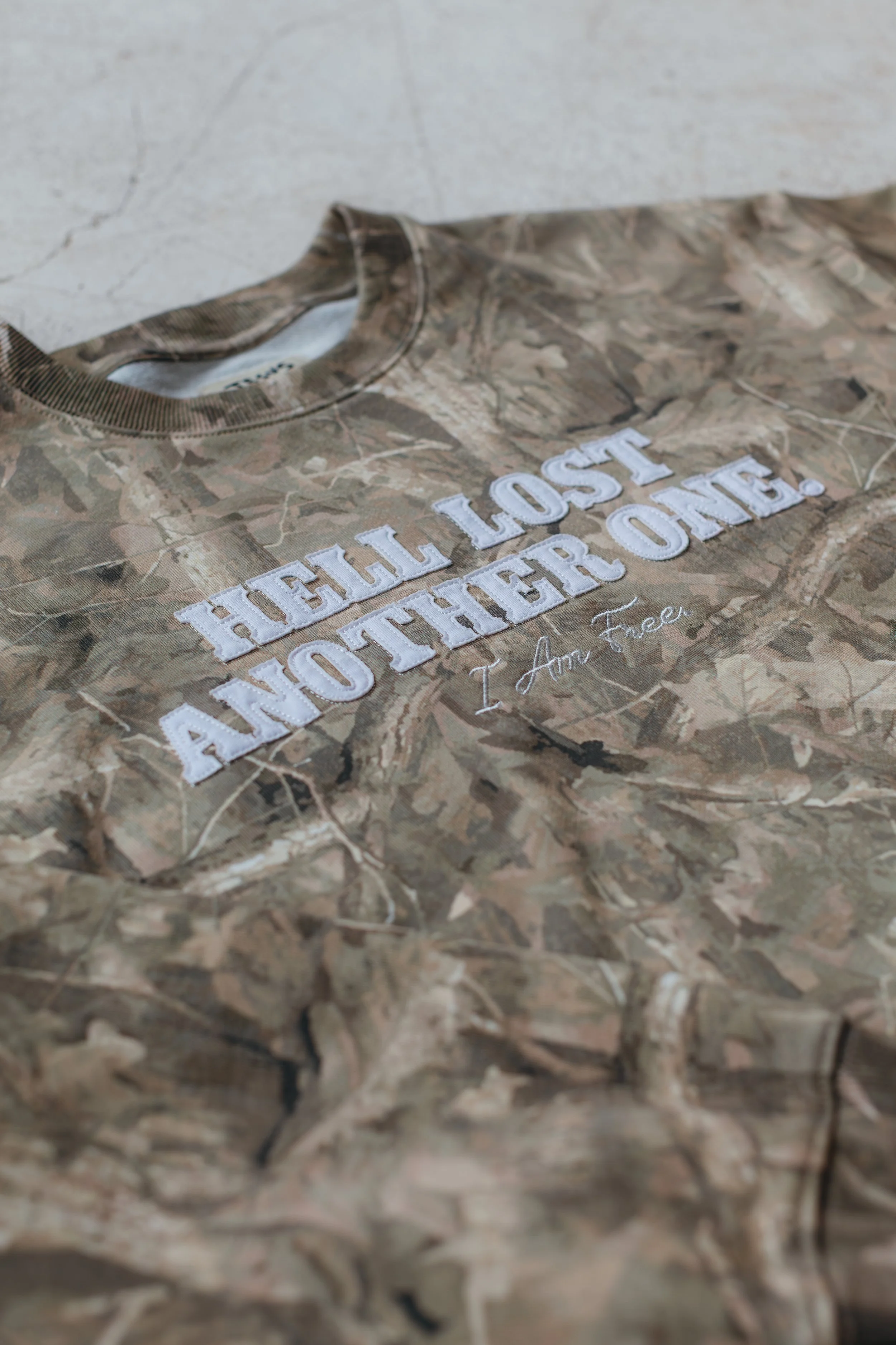 A camo-patterned T-shirt with embroidered text reading 'HELL LOST ANOTHER ONE. I Am Free.'