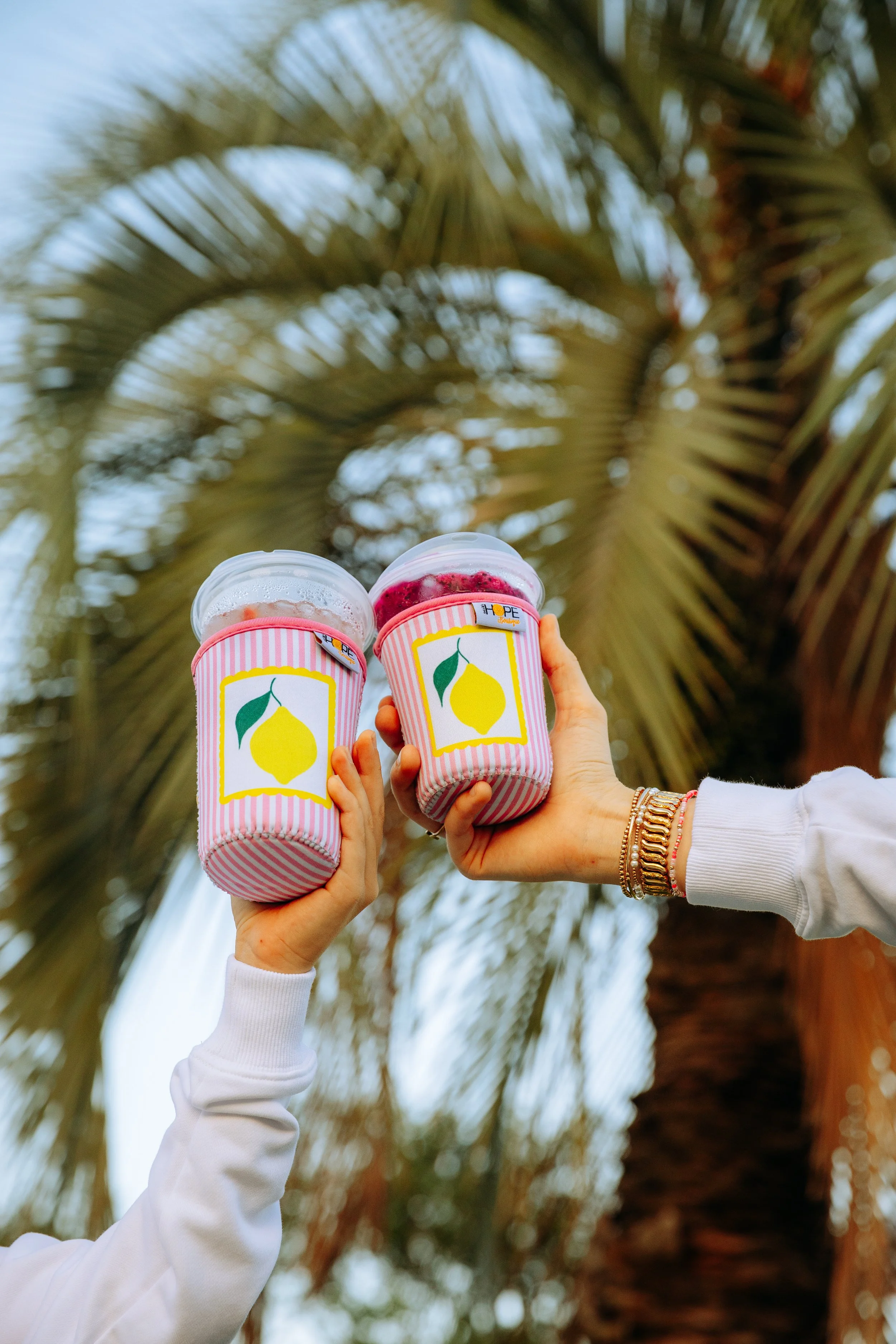 Two hands holding pink striped cups with lemon logos, outdoors with palm trees in background.