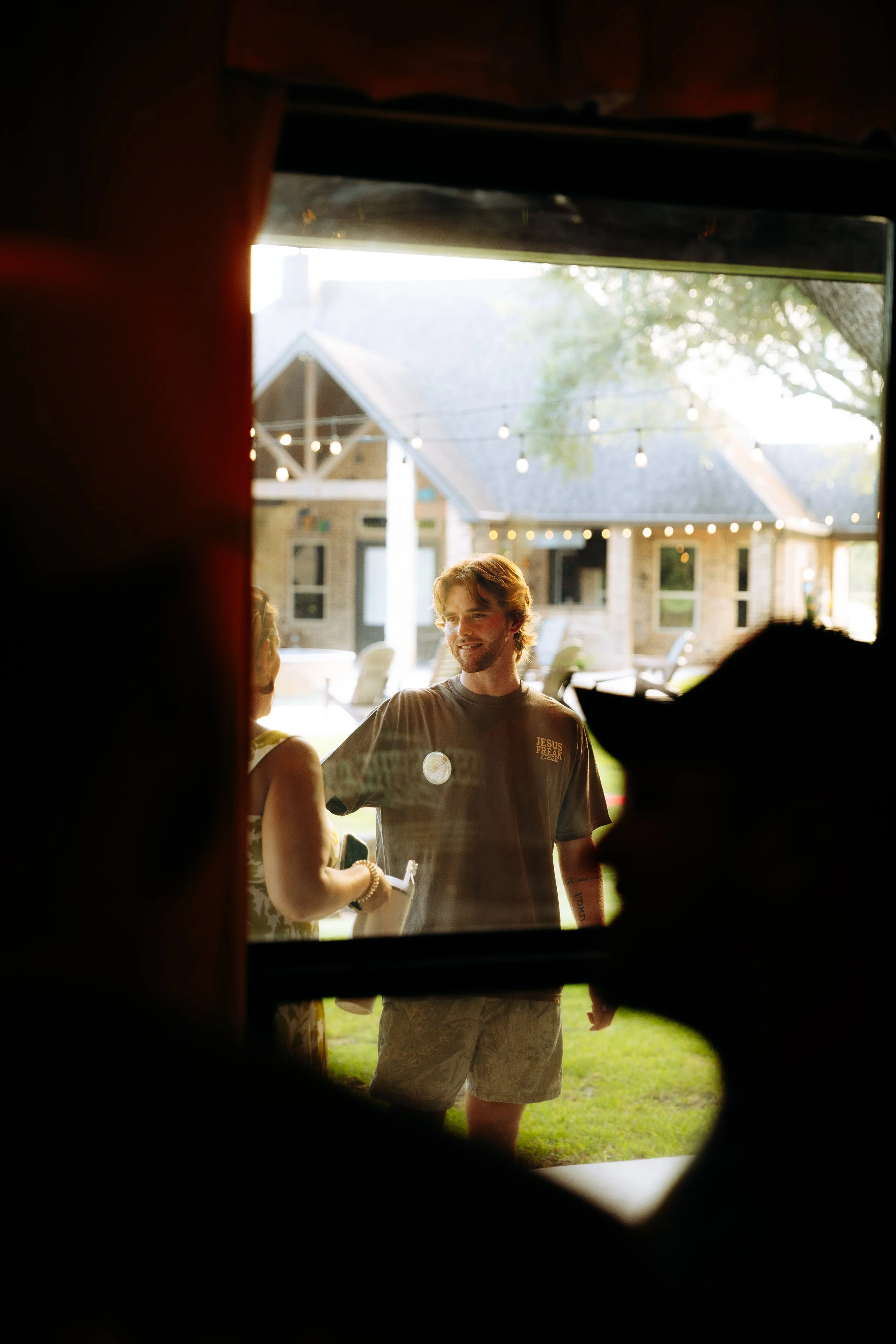 Young man talking to a woman outside a house during a sunny day, viewed through a partially opened window or door.