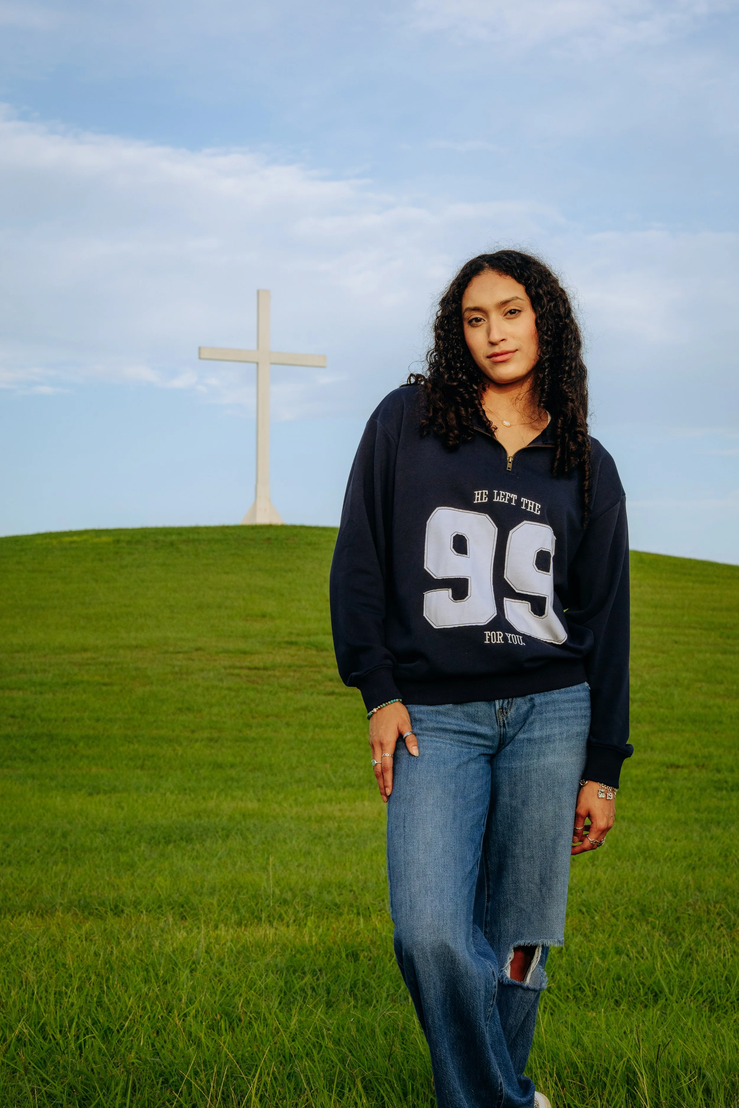 A young woman with curly hair standing on a grassy hill with a white cross in the background, under a partly cloudy sky.