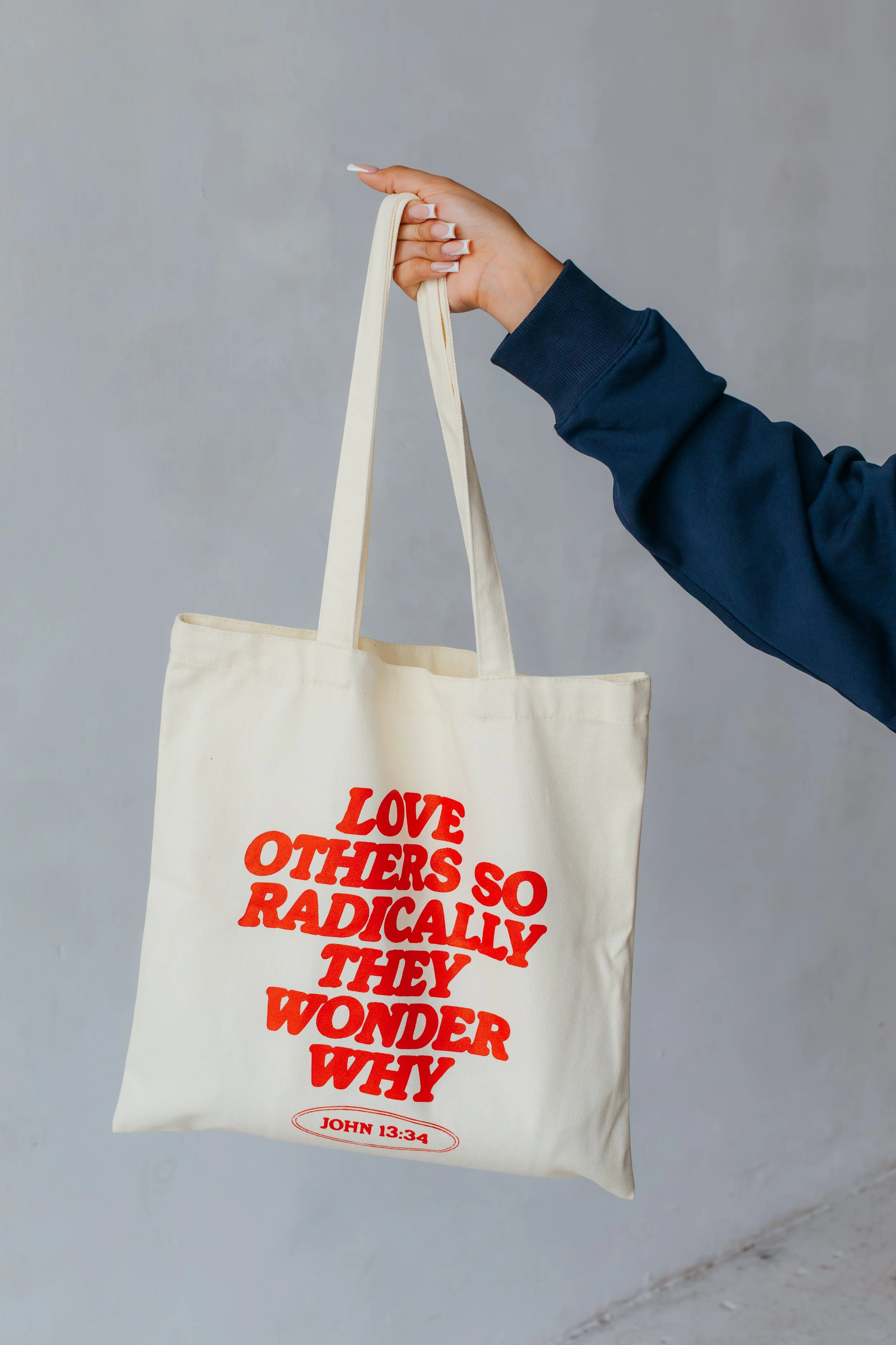 A person holding a beige tote bag with red lettering that reads "Love Others So Radically They Wonder Why" and a Bible reference "John 13:34". The person is wearing a dark blue sleeve and has manicured nails.
